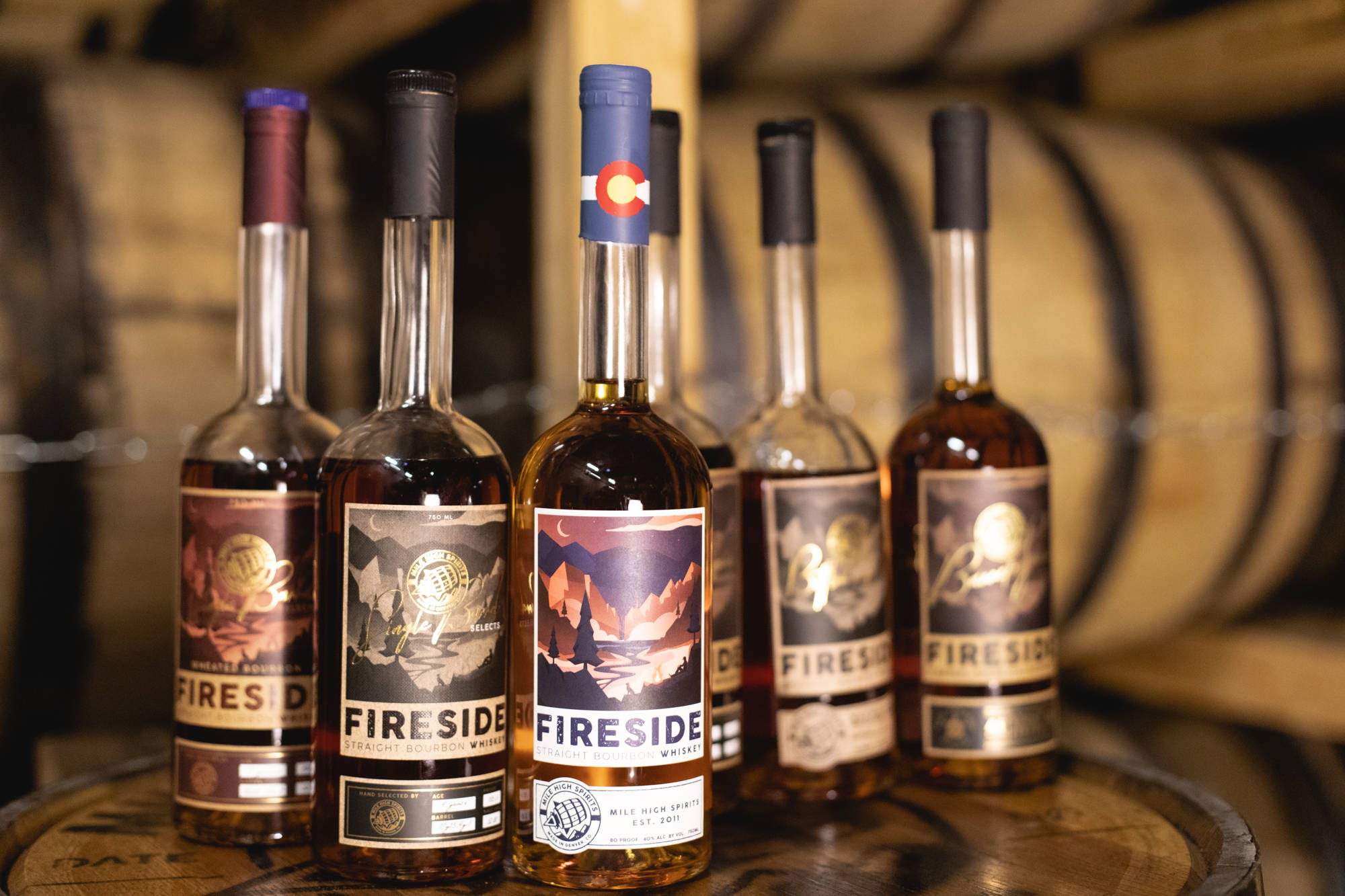 alcohol 桌布 壁紙 — 五瓶帶有山脈標籤的Fireside威士忌酒瓶放在一個木桶上，背景是模糊的威士忌酒桶，位於質樸的釀酒廠。