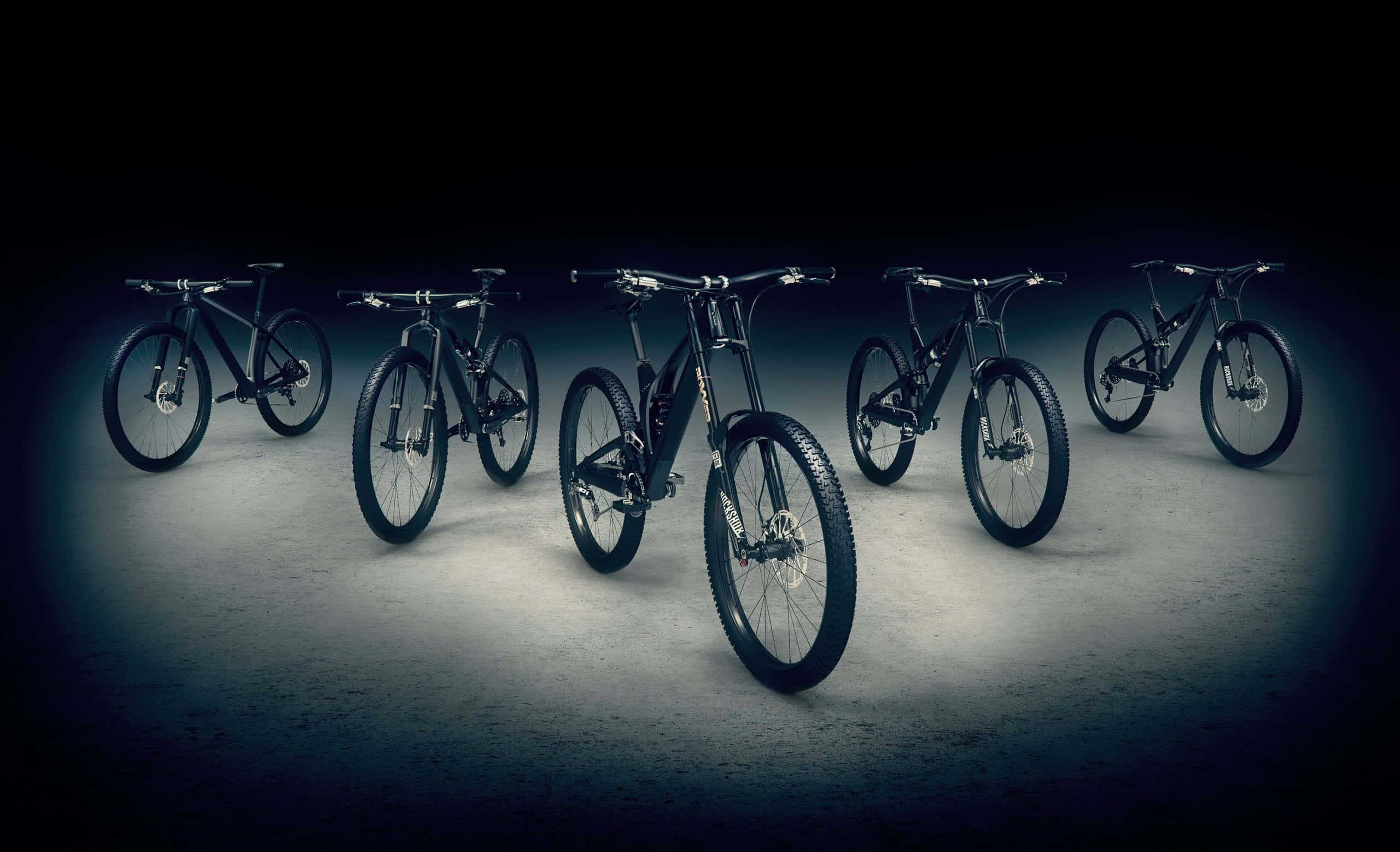 Best Bikes 桌布 壁紙 — 五輛時尚的黑色登山車在黑暗的攝影棚中，以聚光燈效果戲劇性地照亮在混凝土地板上。