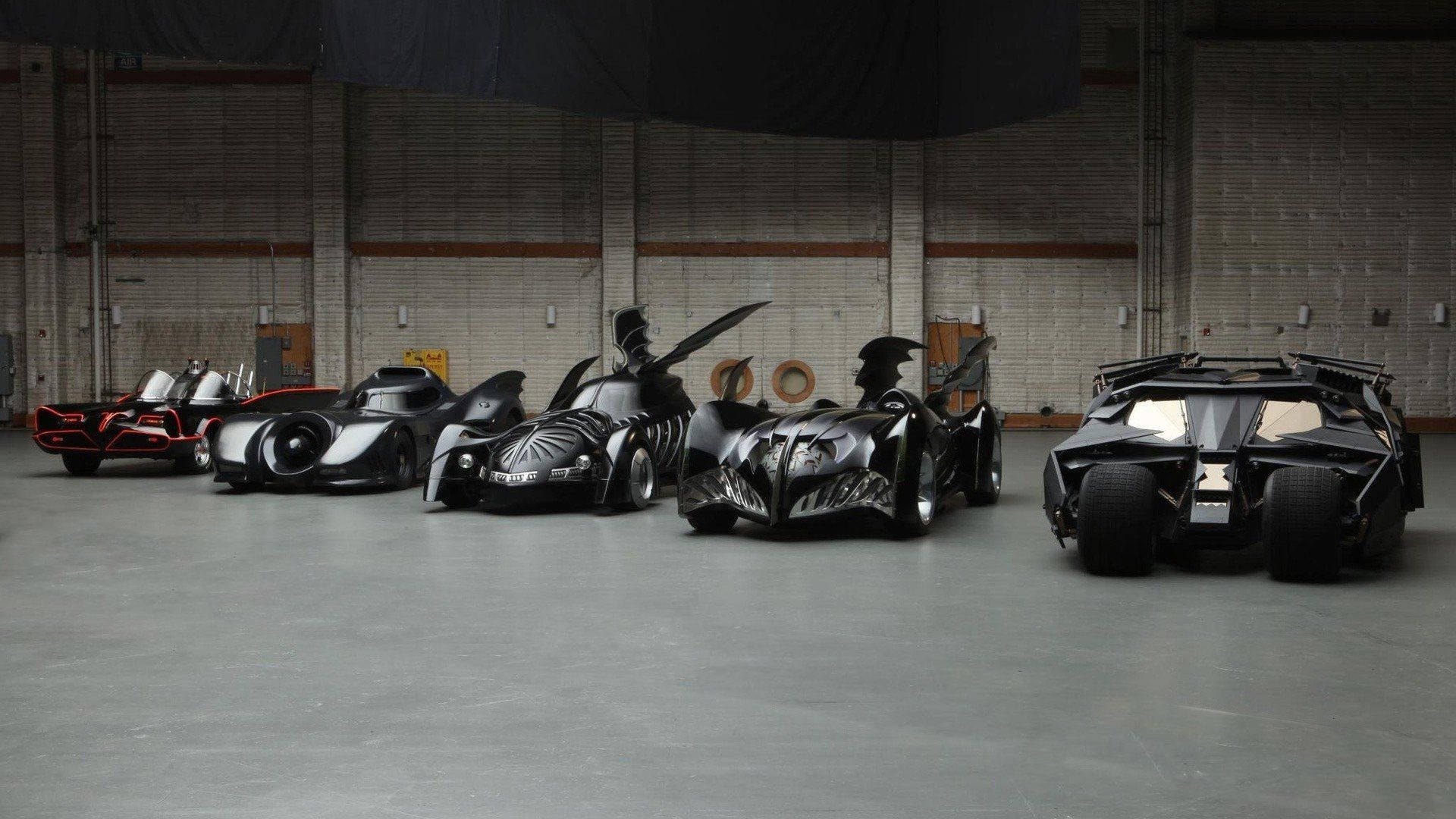 Batmobile 桌布 壁紙 — 五輛標誌性蝙蝠車排列在光線昏暗的工業車庫中，車庫有混凝土牆壁和地板。