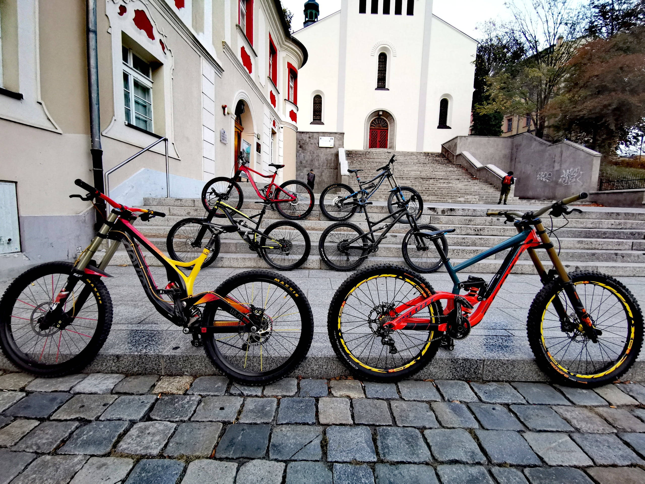 Mountain Bike 桌布 壁紙 — 五輛色彩繽紛的登山自行車停放在白色教堂前的石階上，教堂有紅色點綴和秋天的樹木。