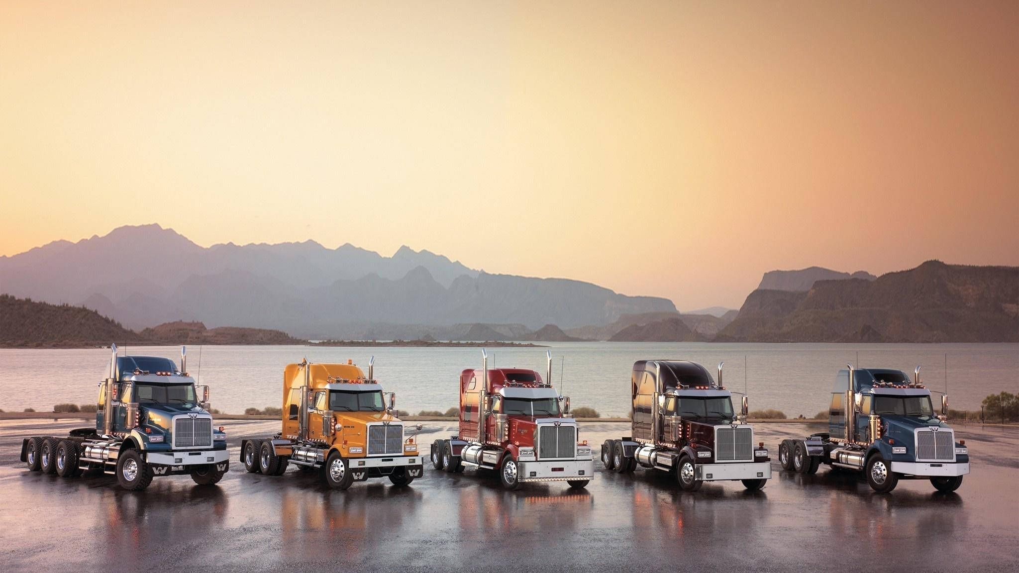 Trucks 桌布 壁紙 — 五輛色彩繽紛的貨櫃車在日落時分排列停在濕滑路面上，背後有山脈和水域。