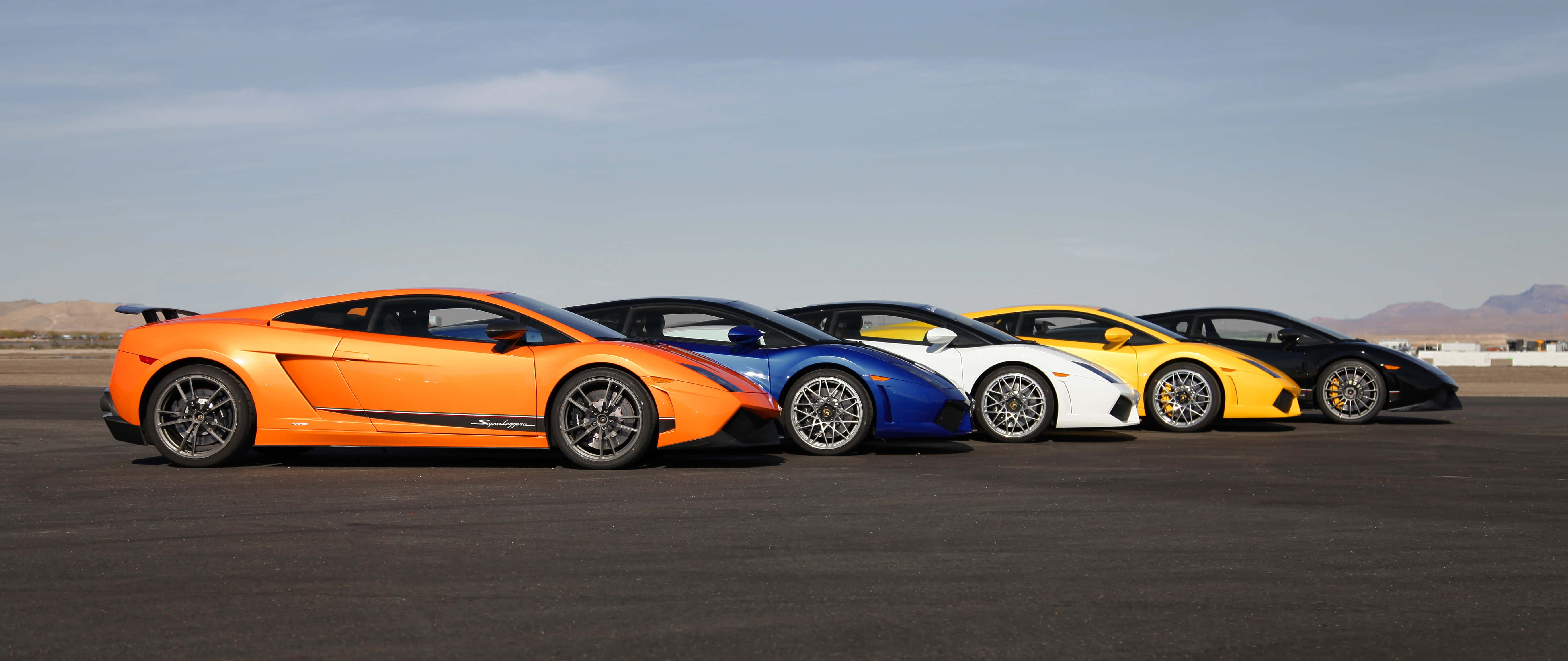 Car Racing 桌布 壁紙 — 五輛色彩鮮豔的藍寶堅尼Gallardo，橙色、藍色、白色、黃色和黑色，在晴朗的天空下排列在沙漠賽道上。