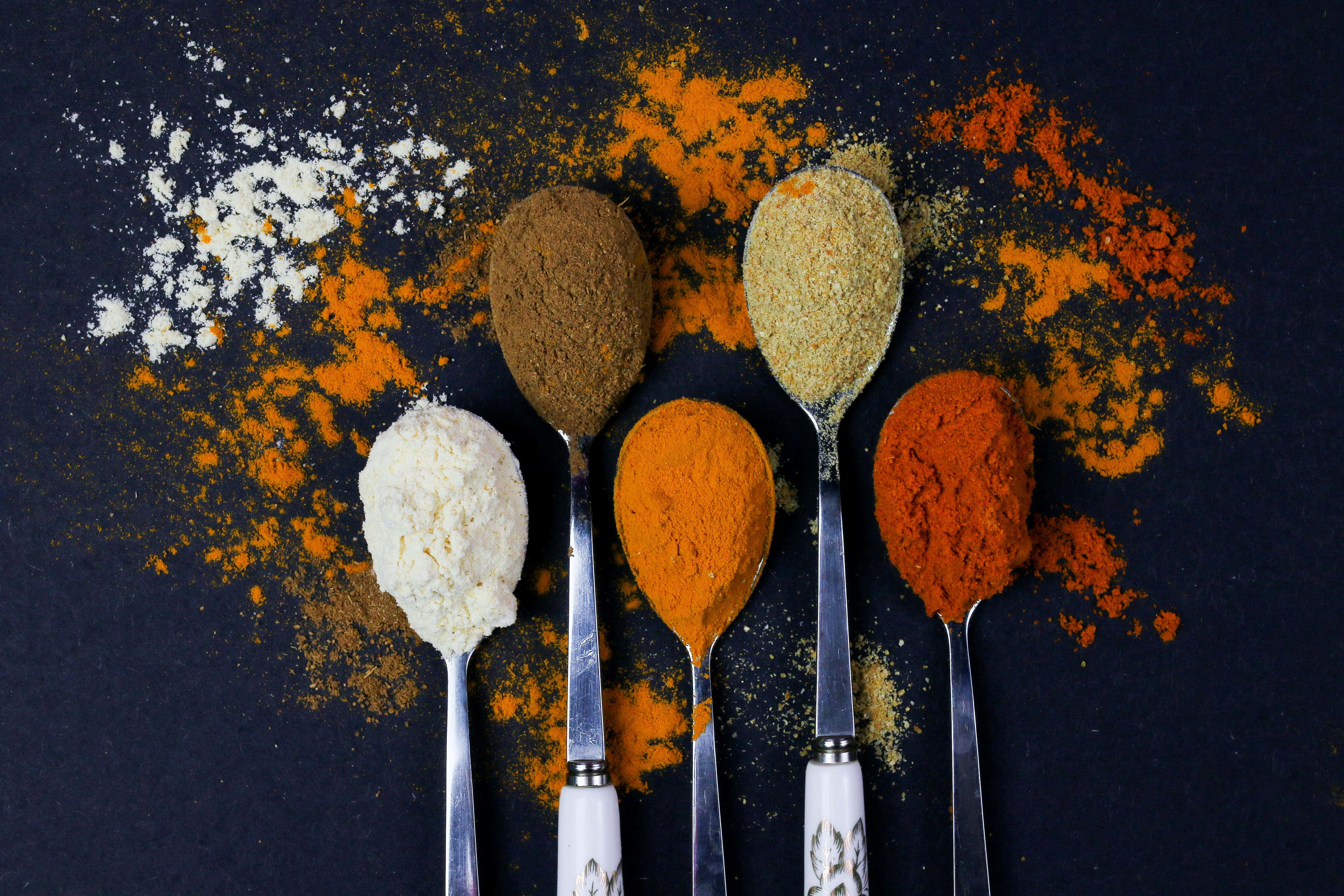 Spices 桌布 壁紙 — 五隻湯匙盛滿了充滿活力的香料，如薑黃和小茴香，放在一個有粉末散落的深色表面上。