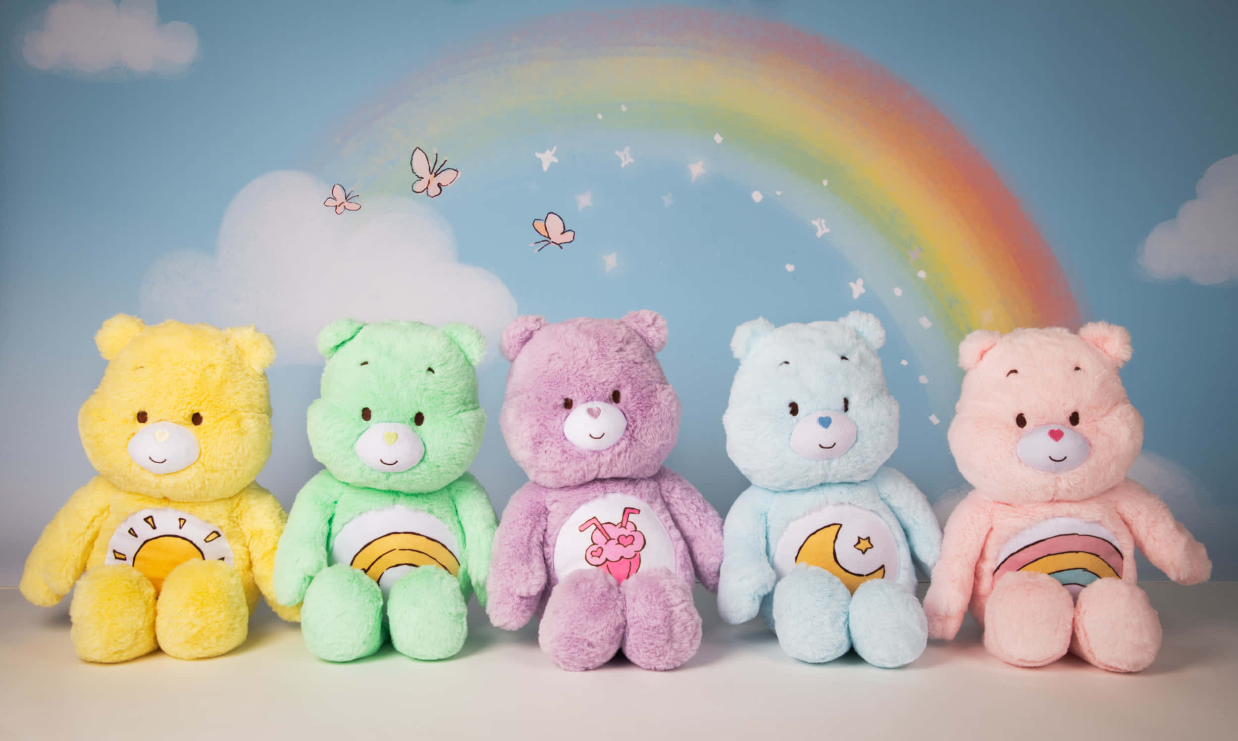 彩虹熊 桌布 壁紙 — 五隻色彩繽紛的Care Bears毛絨玩具坐在畫有彩虹、蝴蝶和雲朵的圖案下一字排開。
