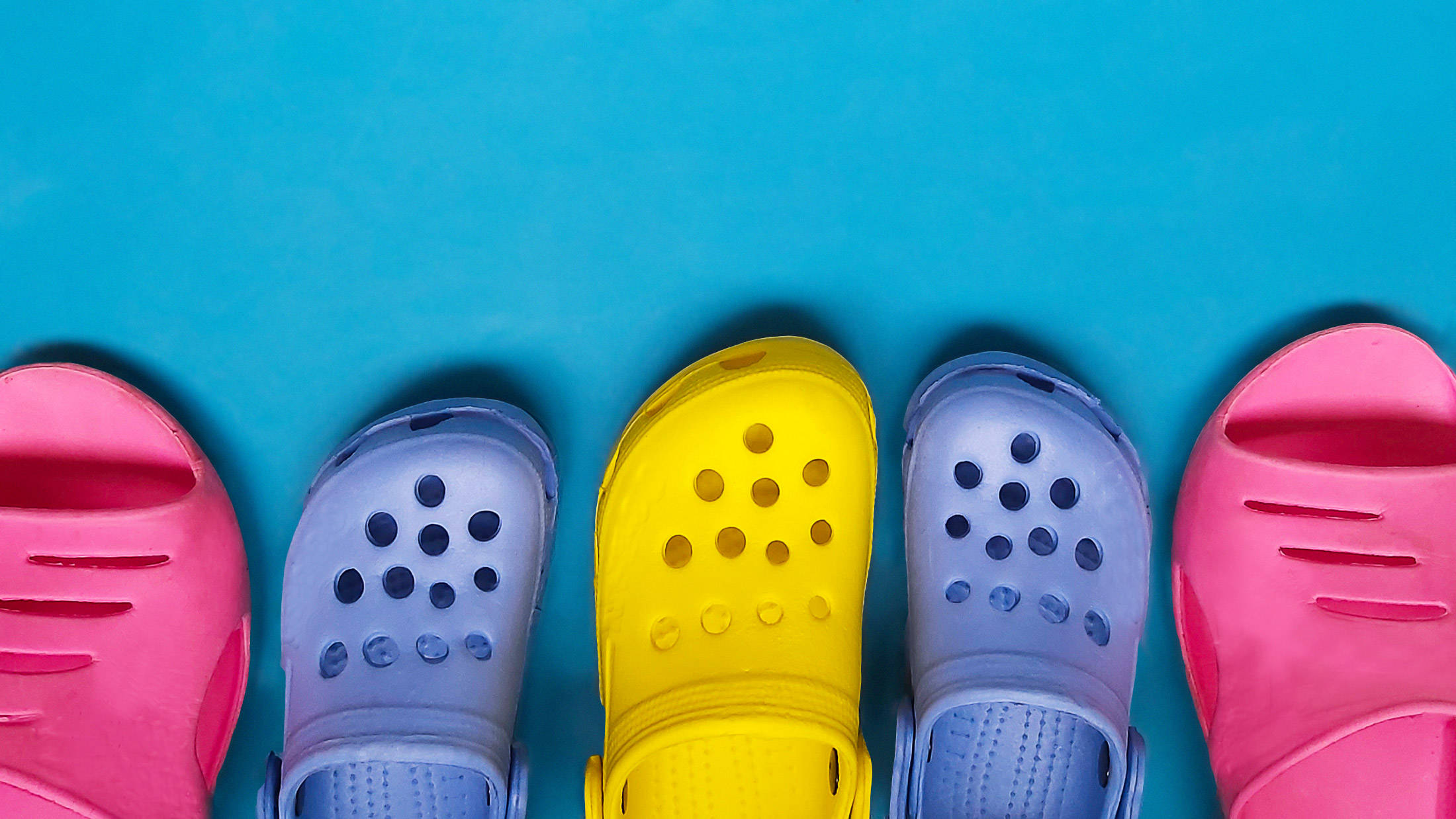 Crocs 桌布 壁紙 — 五雙色彩繽紛的克洛格涼鞋，粉紅色、藍色和黃色，整齊地排列在明亮的藍色背景上。