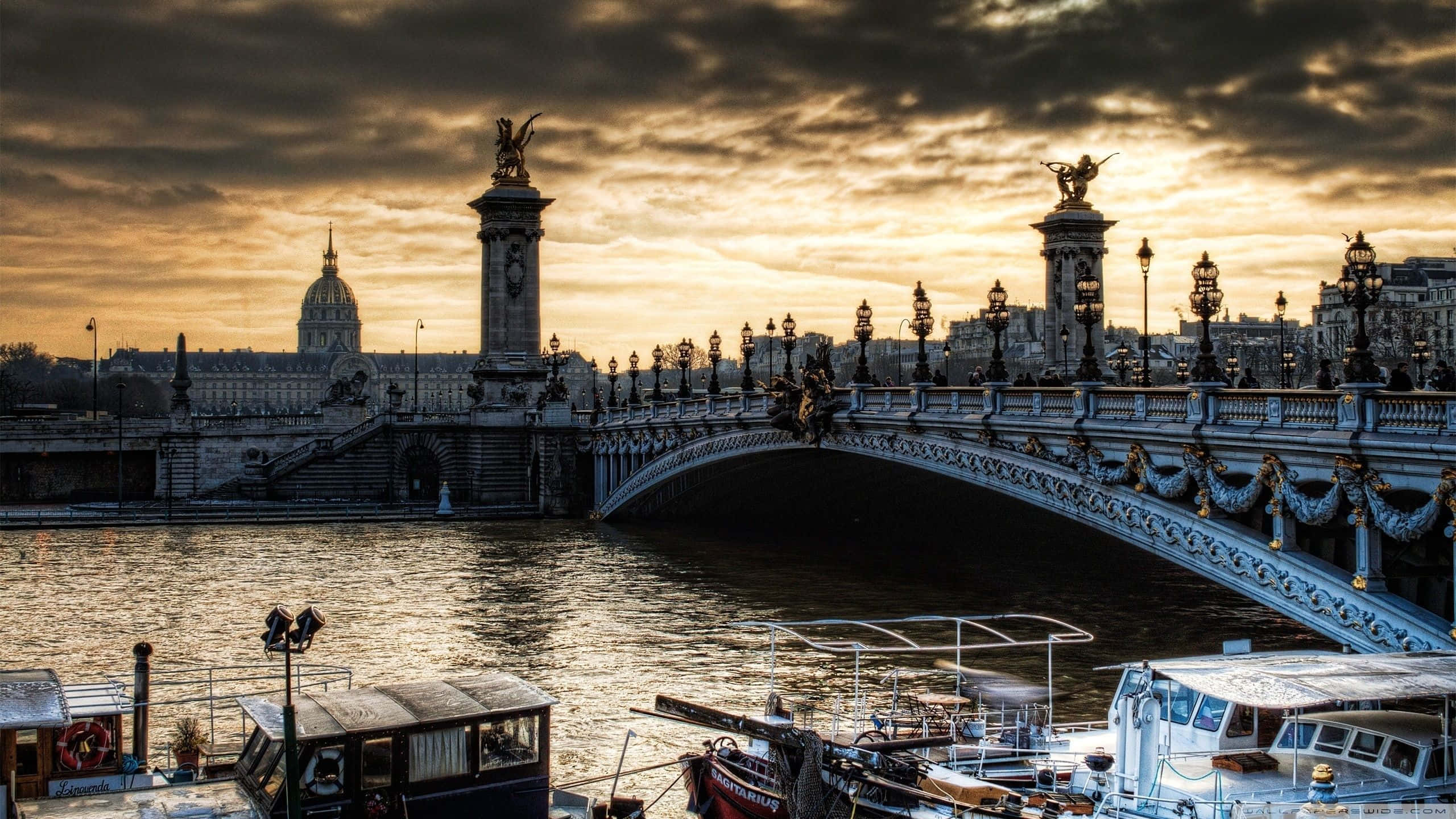 Paris Desktop 桌布 壁紙 — 亞歷山大三世橋在塞納河上日落時閃耀金光，下方停泊著船隻，背景是巴黎建築。