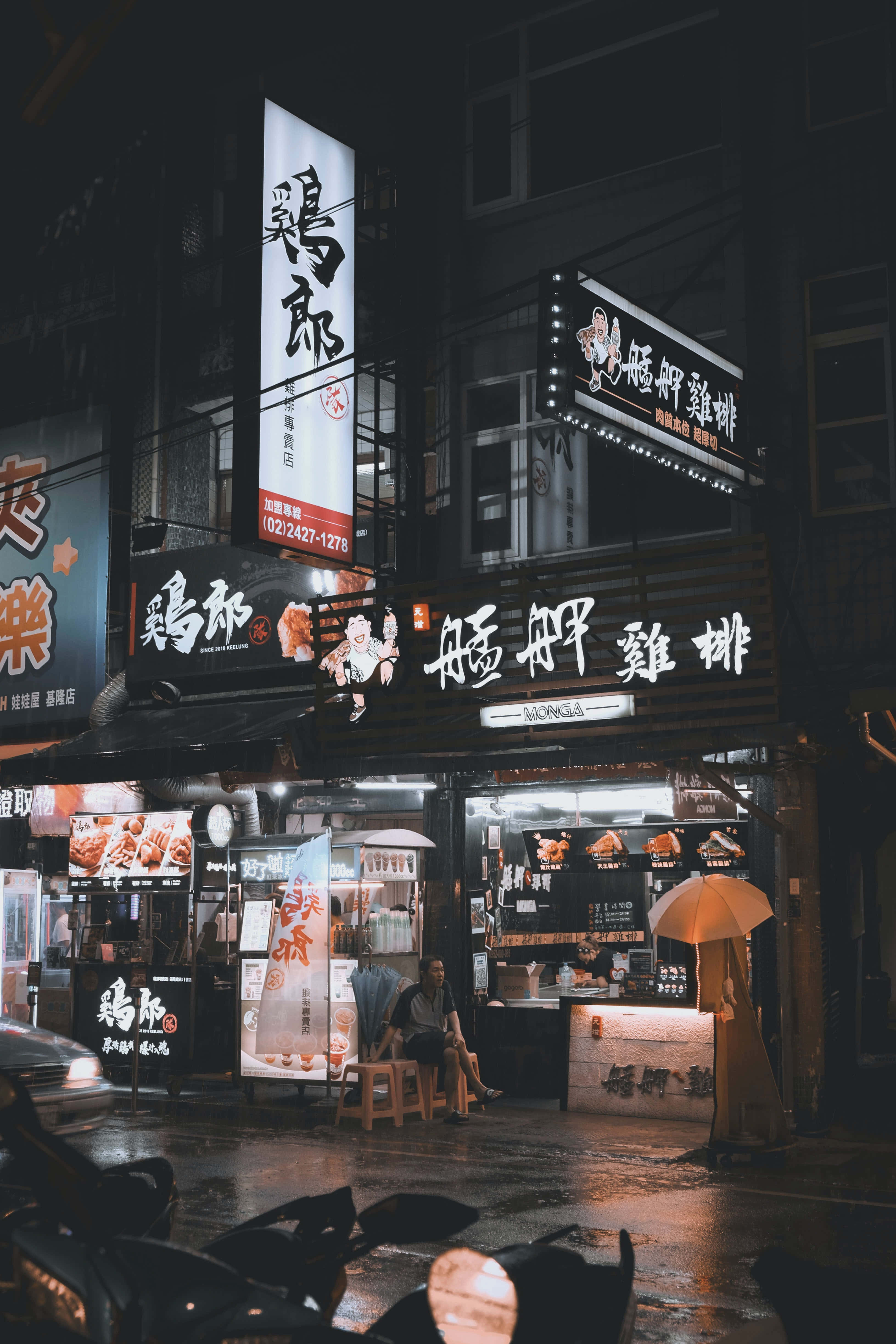 Shop 桌布 壁紙 — 亞洲城市雨夜的街景，發光的小吃攤招牌和一個坐在雨傘下的人。