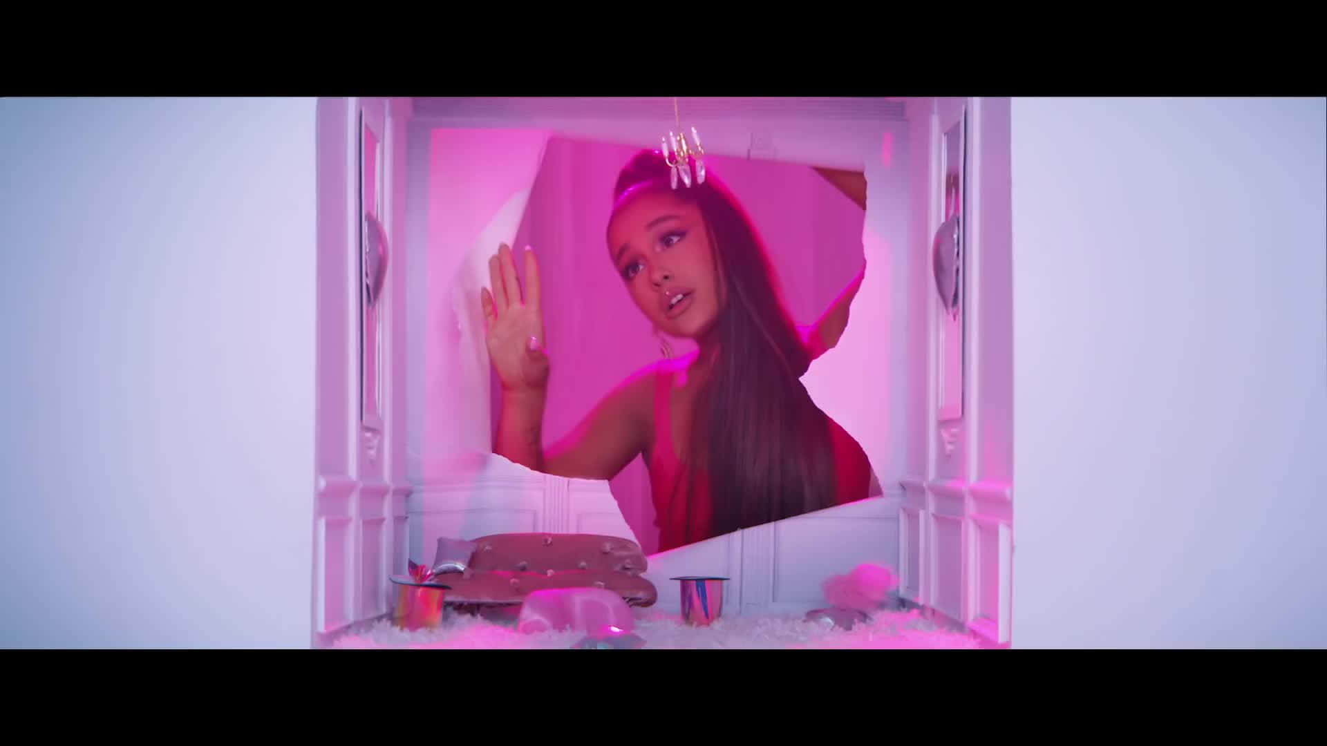 Ariana Grande 7 Rings 桌布 壁紙 — 亞莉安娜·格蘭德在一個鋪滿白色絨毛地板、超現實的娃娃屋風格房間中，透過一道粉紅色燈光的門口窺視。