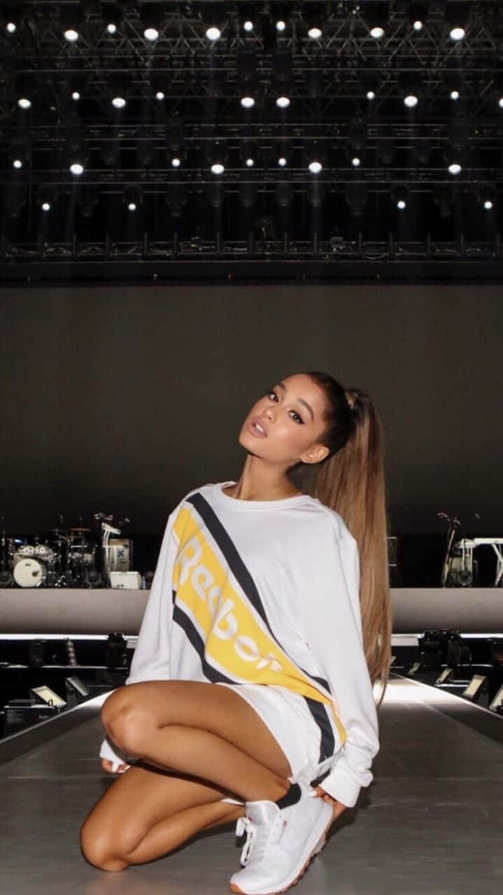 Ariana Grande Aesthetic 桌布 壁紙 — 亞莉安娜·格蘭德在明亮的舞台燈光下，穿著白色Reebok運動衫和白色運動鞋，自信地蹲在音樂會舞台上。