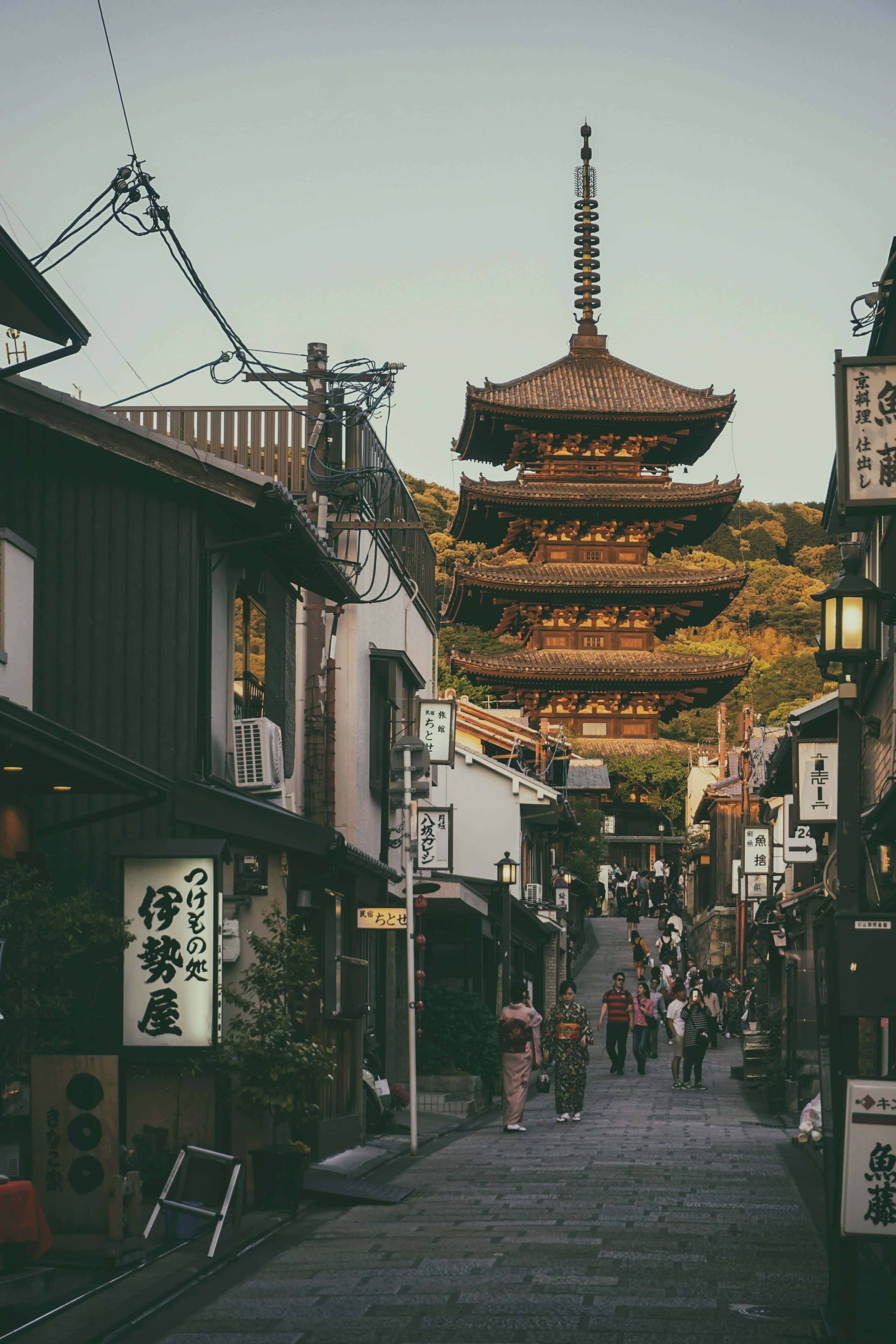 日本 桌布 壁紙 — 京都一條狹窄的石板路，兩旁是傳統木造建築，向上通往一座沐浴在溫暖夕陽中的高聳寶塔。