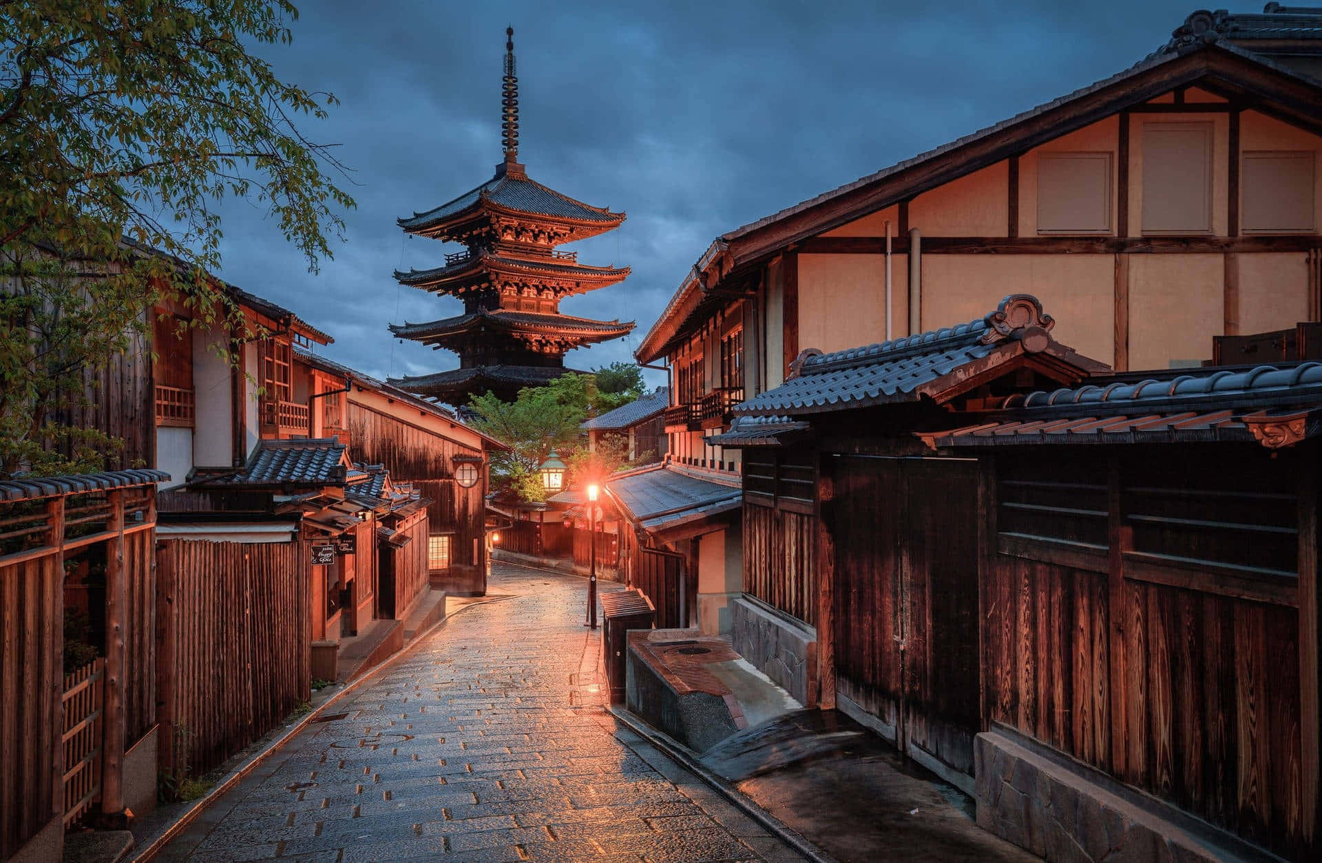 日本 桌布 壁紙 — 京都黃昏時分一條狹窄潮濕的石板小巷，兩旁是木造建築，背景中一座寶塔在陰沉天空下聳立。