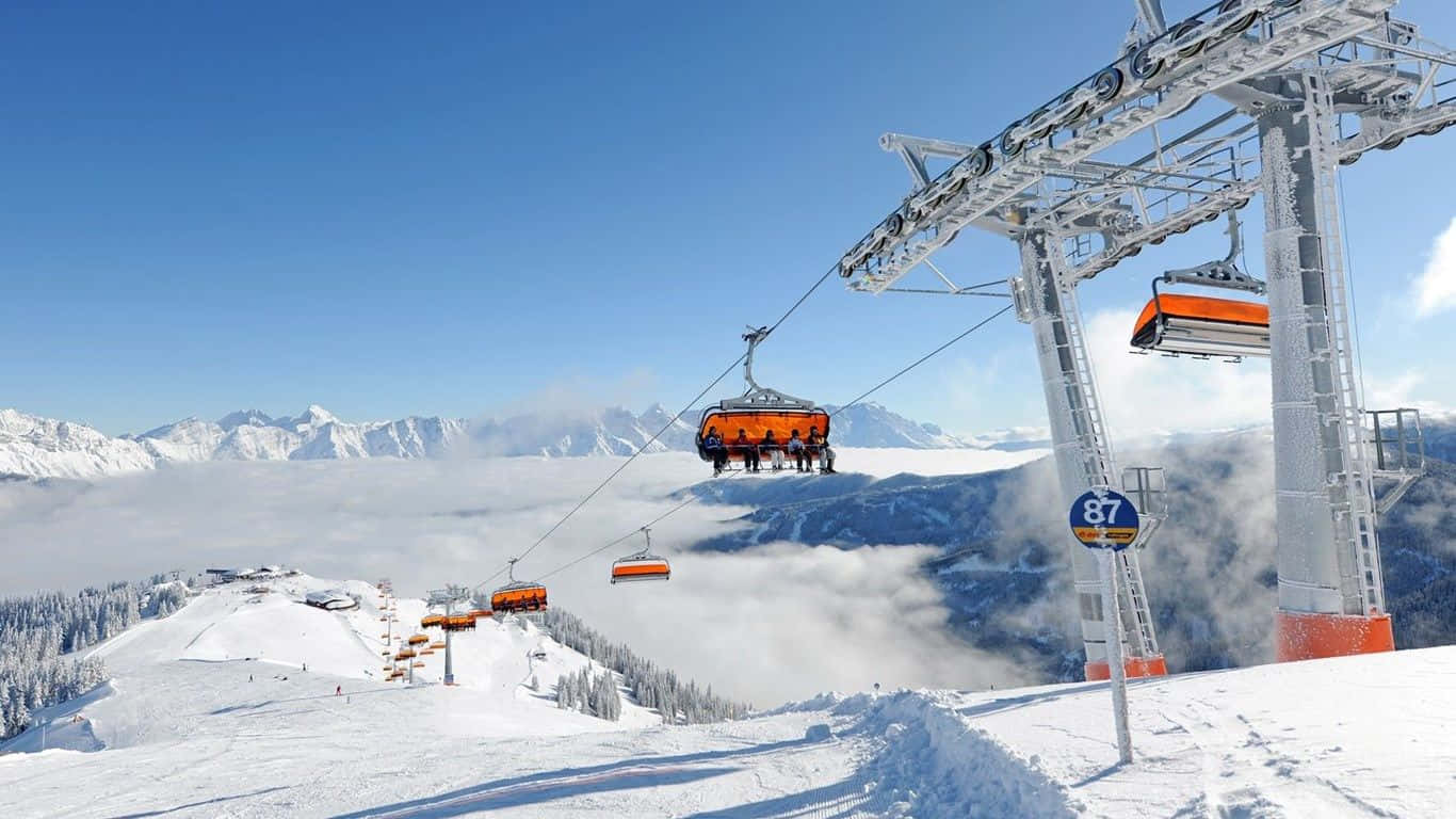 Ski Resort 桌布 壁紙 — 亮橙色滑雪纜車在晴朗藍天下，滑過白雪覆蓋的山峰和雲海之上。