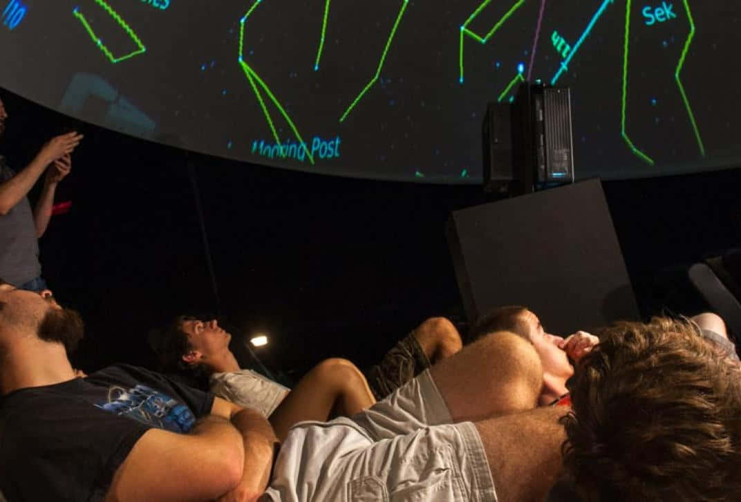 Planetarium 桌布 壁紙 — 人們仰臥著觀看一個大型弧形螢幕，螢幕上顯示發光的綠色星座和星星名稱，房間昏暗。