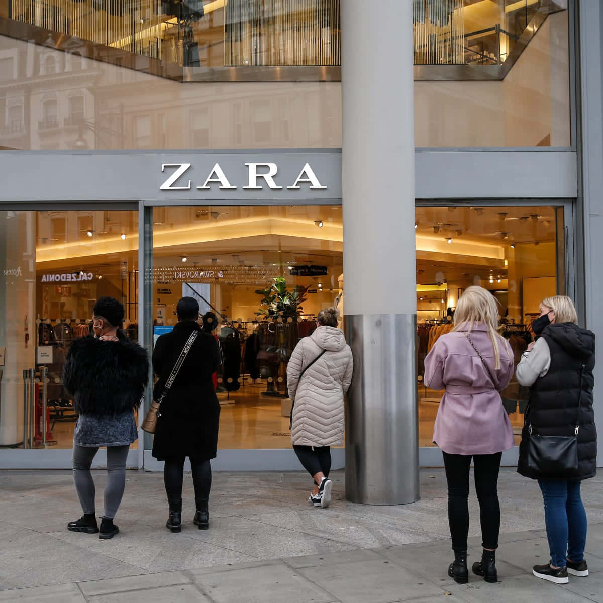 Zara 桌布 壁紙 — 人們在 Zara 商店外排隊，玻璃窗反射城市建築物和溫暖的室內燈光。
