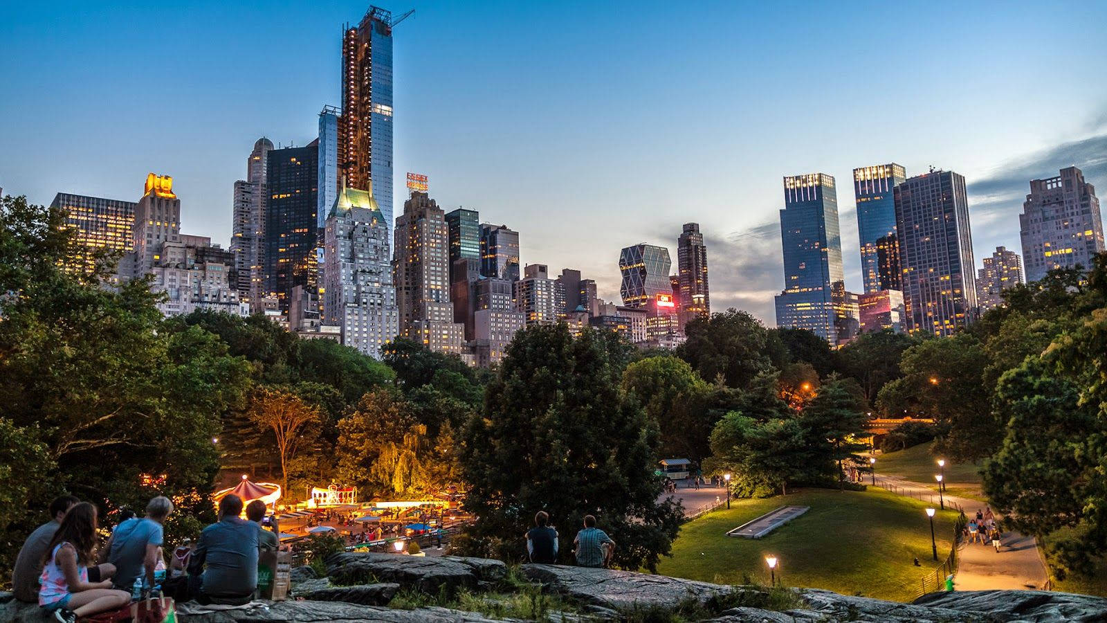 New York Street 桌布 壁紙 — 人們在岩石上放鬆，俯瞰暮色中充滿活力的城市天際線，下方是閃閃發光的摩天大樓和鬱鬱蔥蔥的公園。