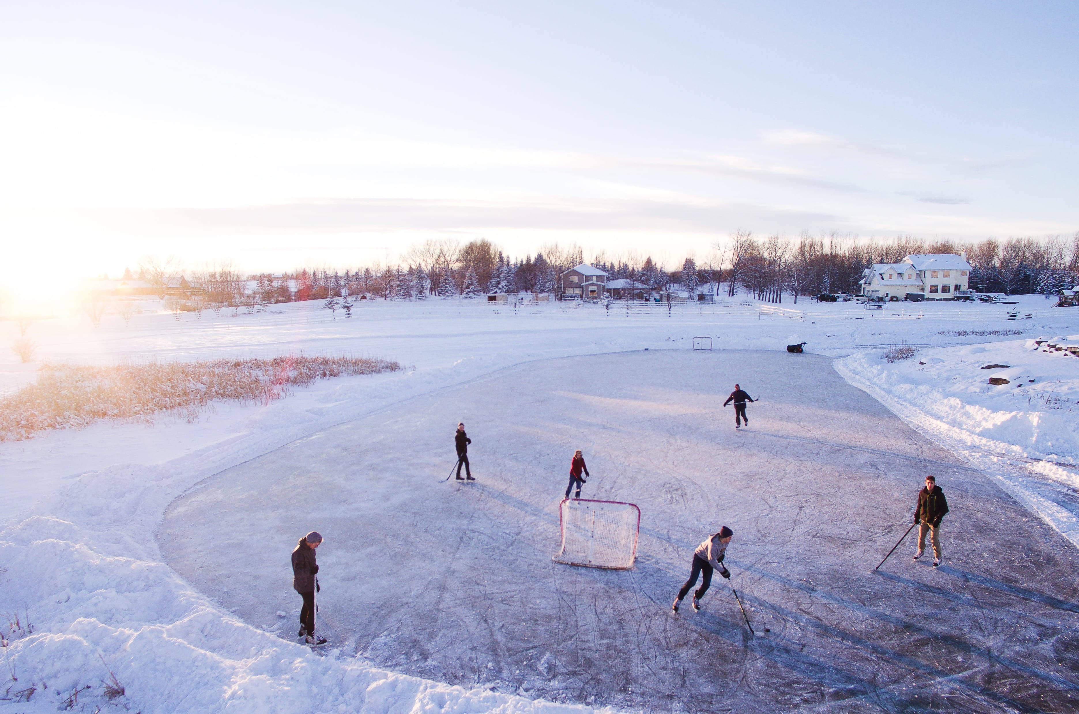 hockey 桌布 壁紙 — 人們在日落時分的冰凍池塘上打曲棍球，背景是積雪覆蓋的房屋和光禿禿的樹木。