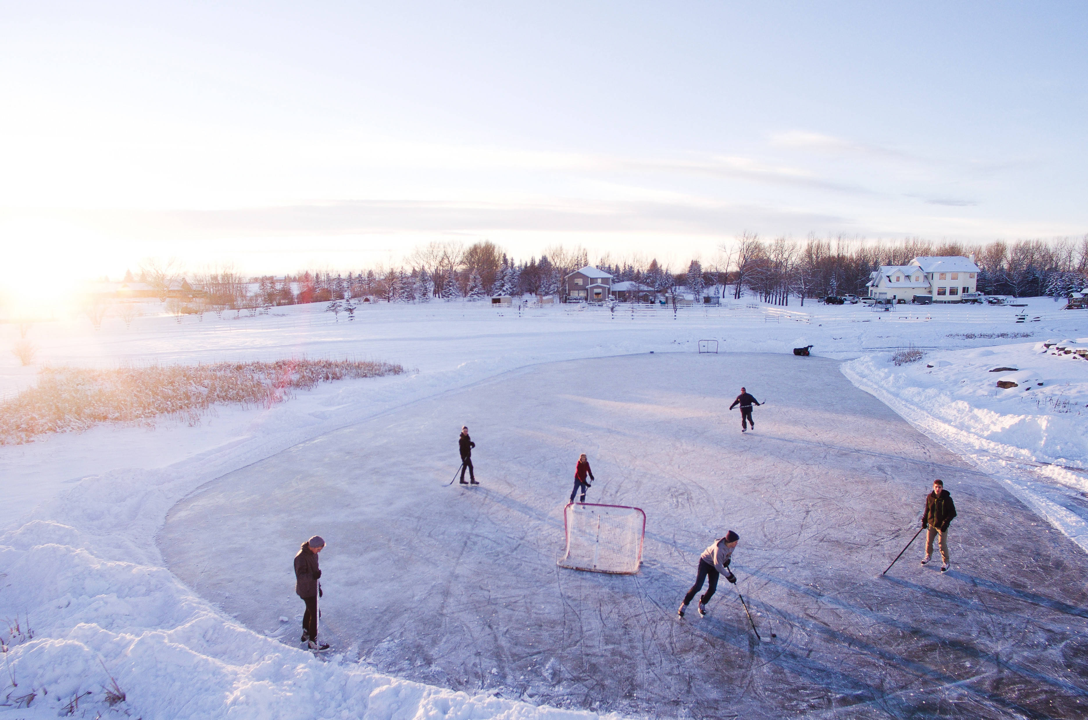 Cool Hockey 桌布 壁紙 — 人們在日落時分的冰凍池塘上打曲棍球，背景是積雪覆蓋的房屋和光禿禿的樹木。