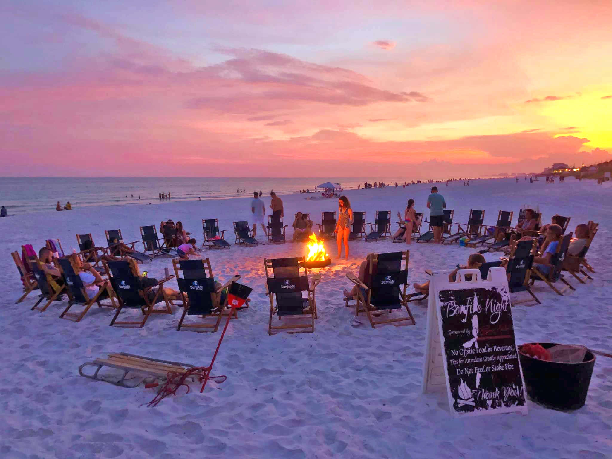 Beach Bonfire 桌布 壁紙 — 人們在日落時分聚集在海灘營火旁，天空呈現粉橙色，海洋地平線清晰可見。