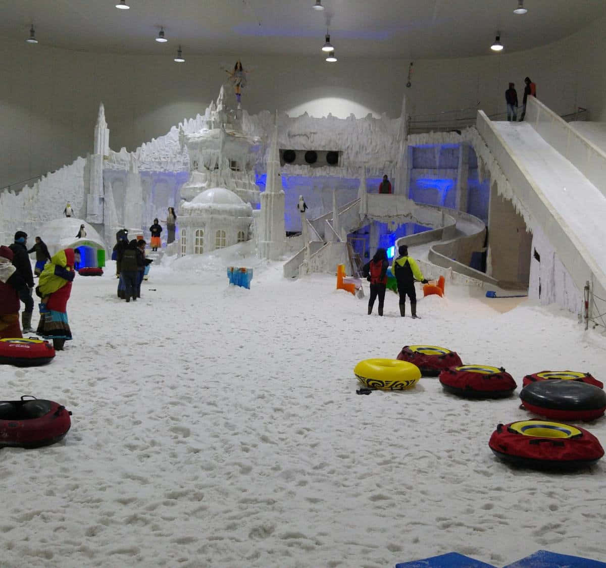 雪 桌布 壁紙 — 人們在明亮照明的室內冬季仙境中享受雪地輪胎和冰雕，並帶有藍色燈光點綴。