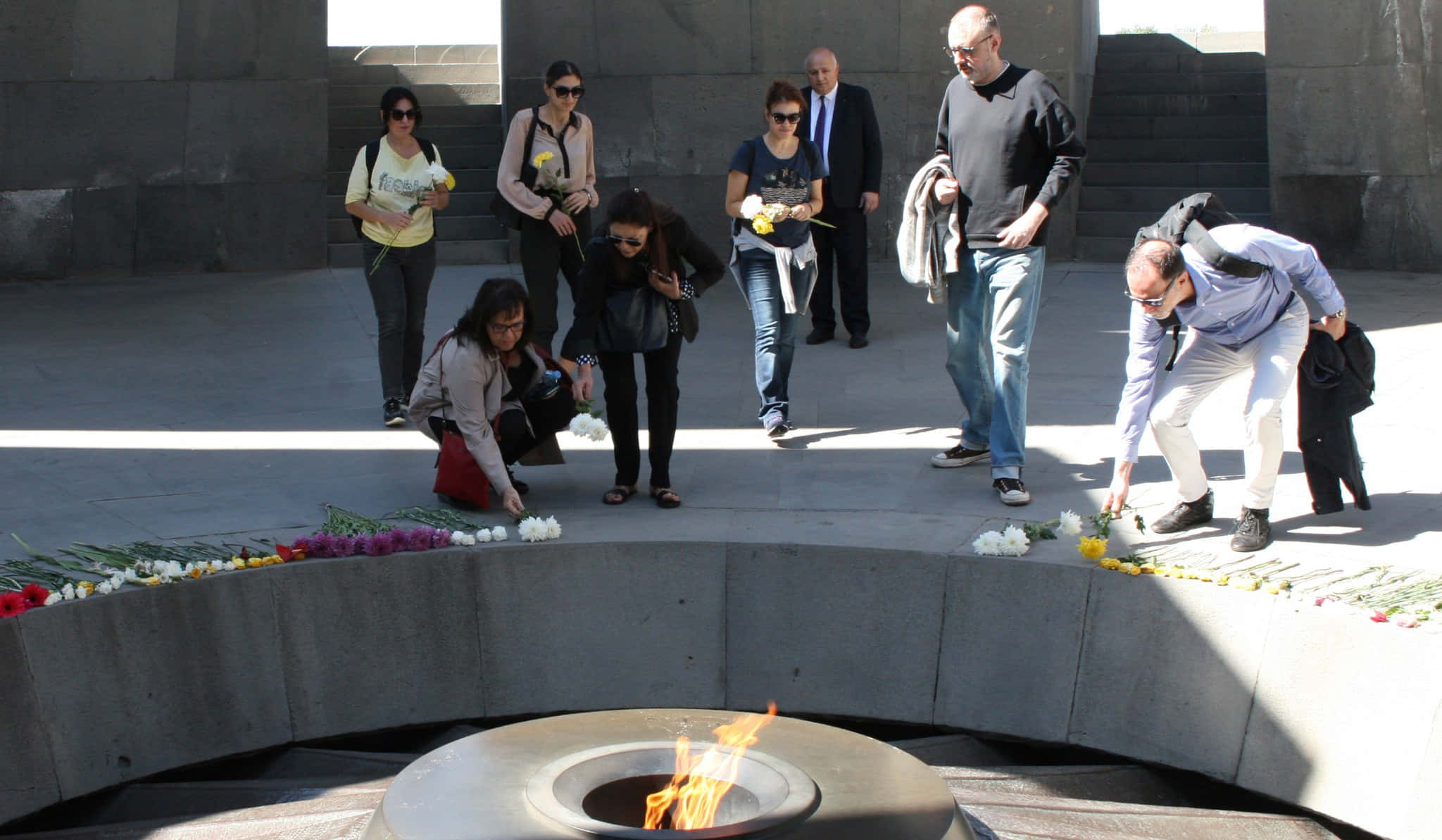 Memorial 桌布 壁紙 — 人們在明亮的陽光下，恭敬地在莊嚴的紀念場所周圍放置鮮花，圍繞著發光的永恆火焰。