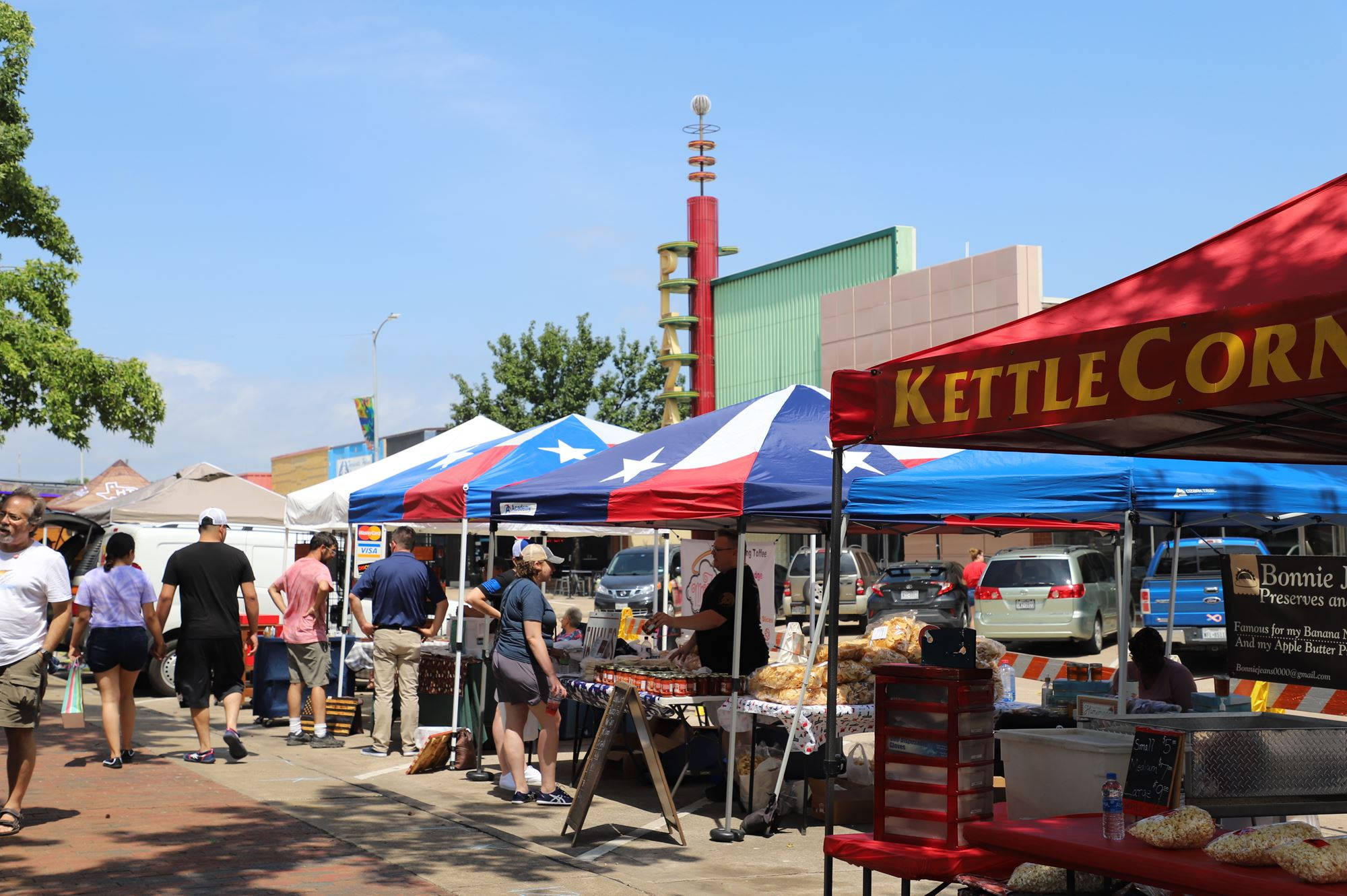 Market 桌布 壁紙 — 人們在晴朗的天空下，在一個充滿活力的戶外市場中瀏覽色彩繽紛的帳篷和爆米花攤位。