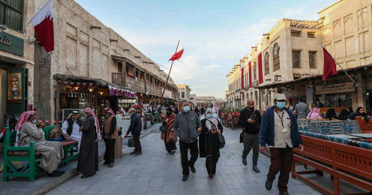 Qatar 桌布 壁紙 — 人們在晴朗的天空下，漫步在一條熙熙攘攘的傳統市場街道上，兩旁是歷史建築和飄揚的國旗。