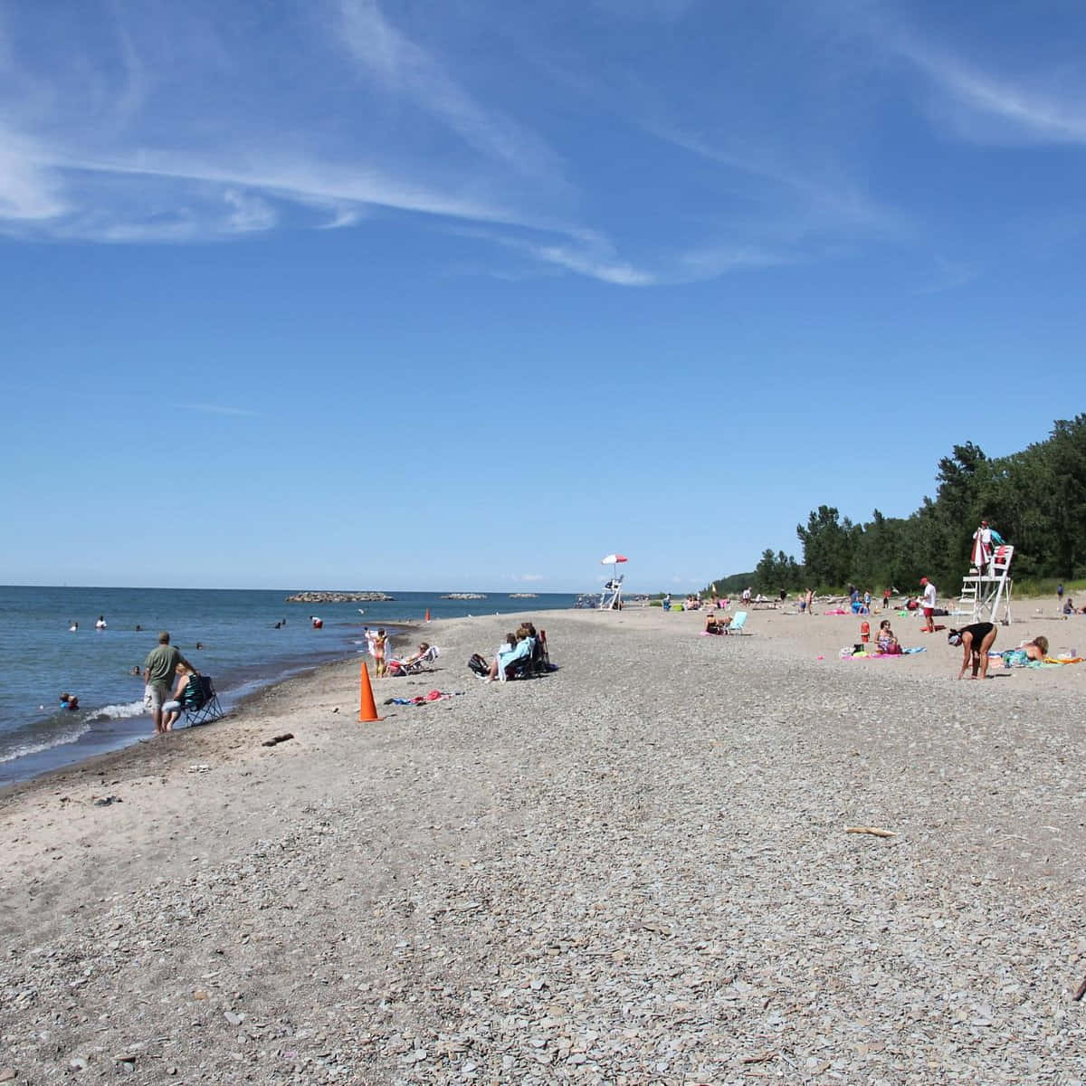 Lake Erie 桌布 壁紙 — 人們在晴朗的日子裡，在有救生塔和遠處樹木的卵石海灘上放鬆和游泳。