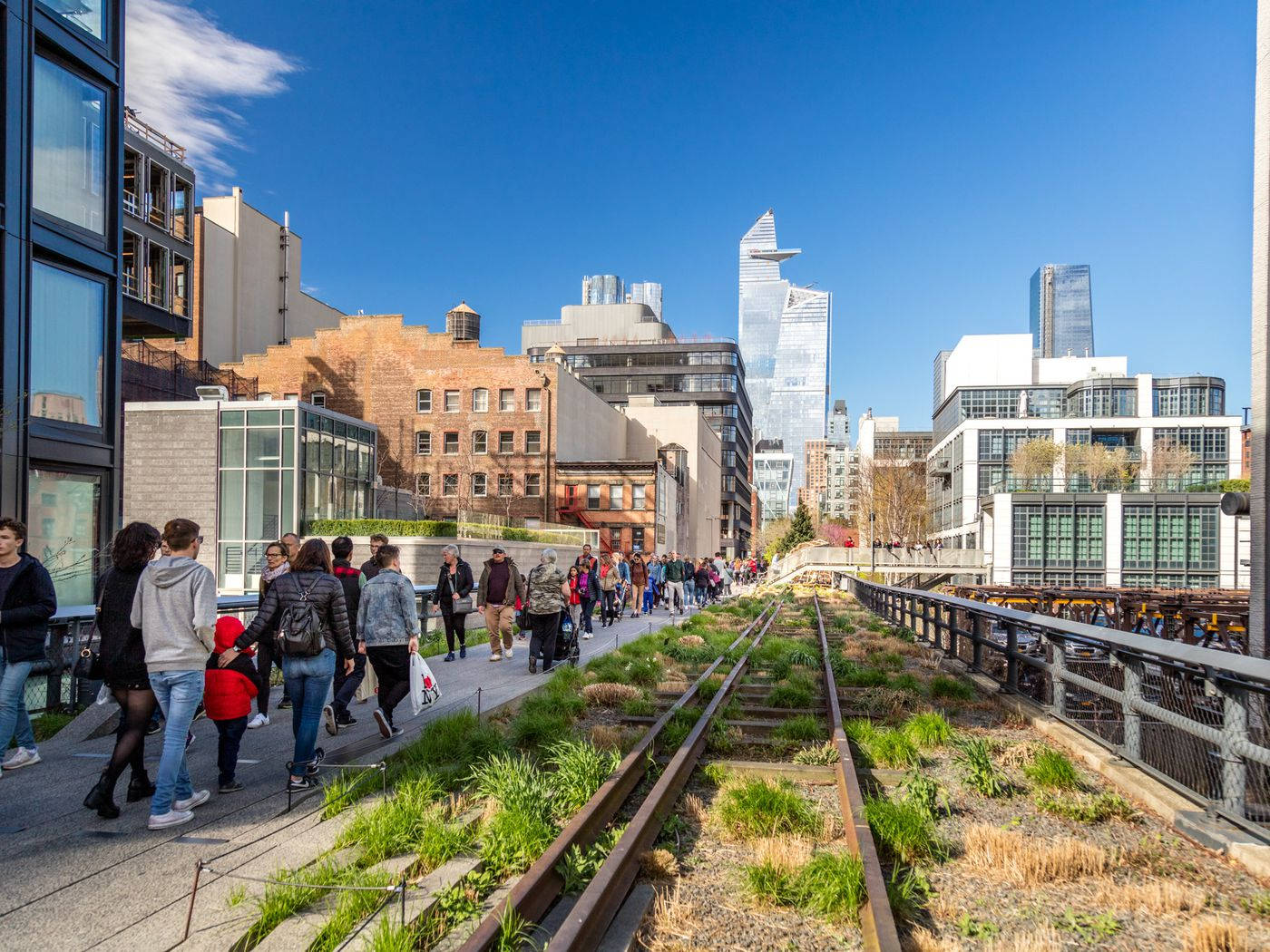 New York 桌布 壁紙 — 人們在晴朗的藍天下，沿著一條充滿活力的城市小徑，旁邊是雜草叢生的火車軌道，背景是現代化的摩天大樓。