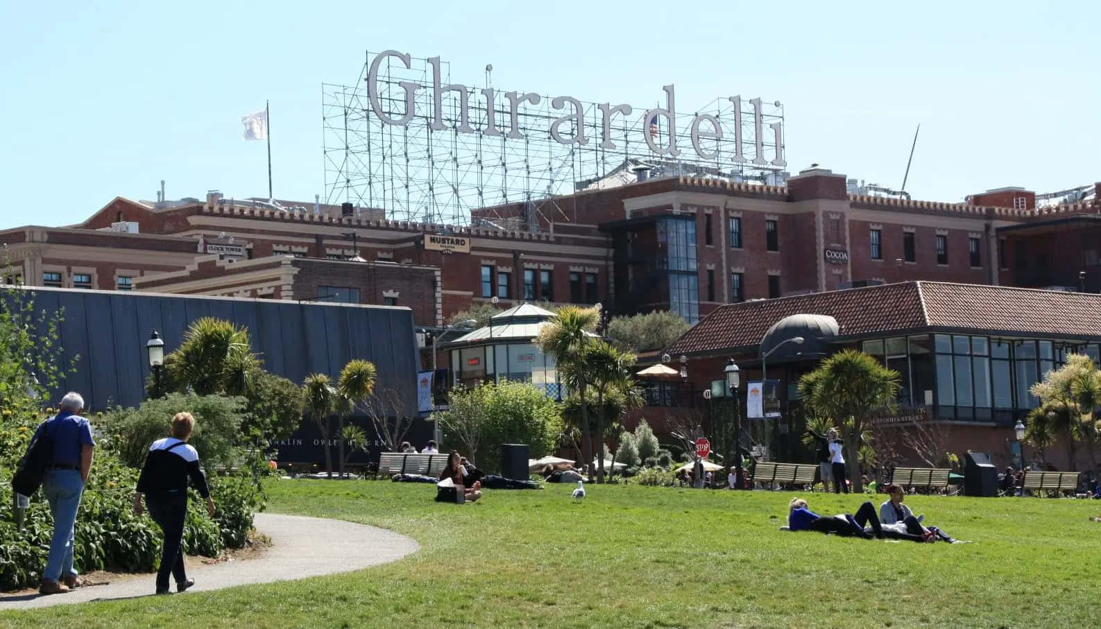 城市 桌布 壁紙 — 人們在晴朗的藍天下在草地上放鬆，標誌性的 Ghirardelli 招牌聳立在磚砌建築和棕櫚樹之上。
