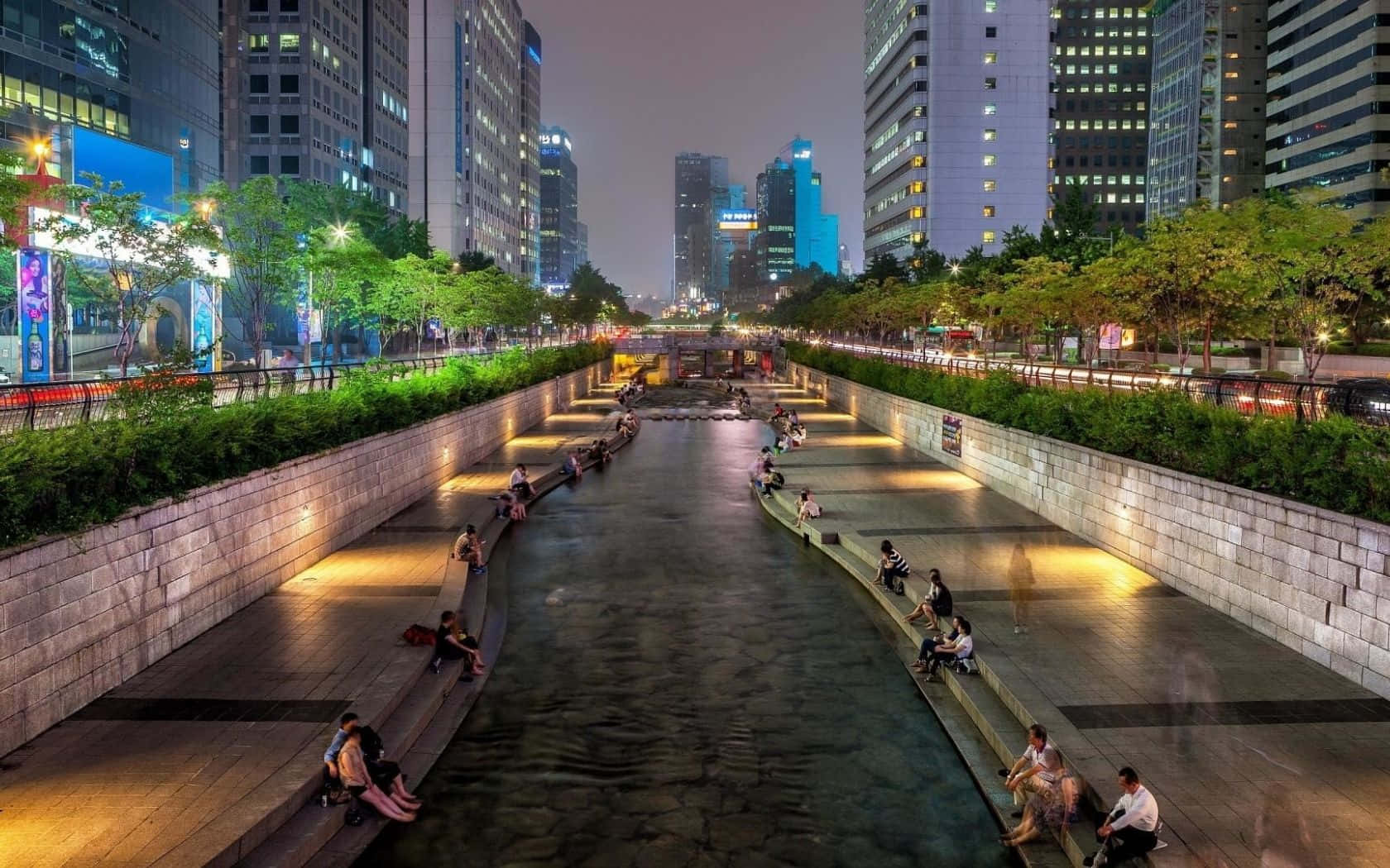 Aesthetic Korean 桌布 壁紙 — 人們在暮色中沿著發光的城市運河放鬆，而城市摩天大樓照亮了他們身後的天際線。