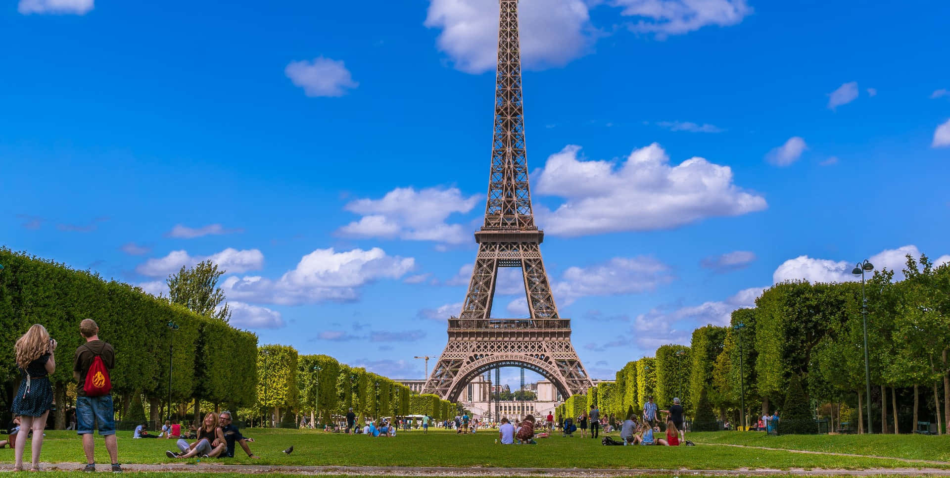 Paris Desktop 桌布 壁紙 — 人們在標誌性的艾菲爾鐵塔附近，在充滿活力的綠色草坪上放鬆和漫步，頭頂是明亮的藍天，點綴著蓬鬆的白雲。