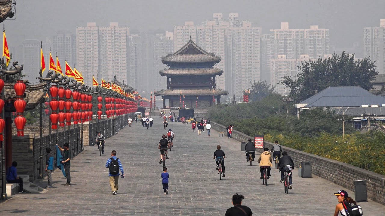 Xian 桌布 壁紙 — 人們在歷史悠久的石牆旁騎腳踏車和散步，石牆上掛滿了紅色燈籠和旗幟，天空朦朧，遠處有現代建築。