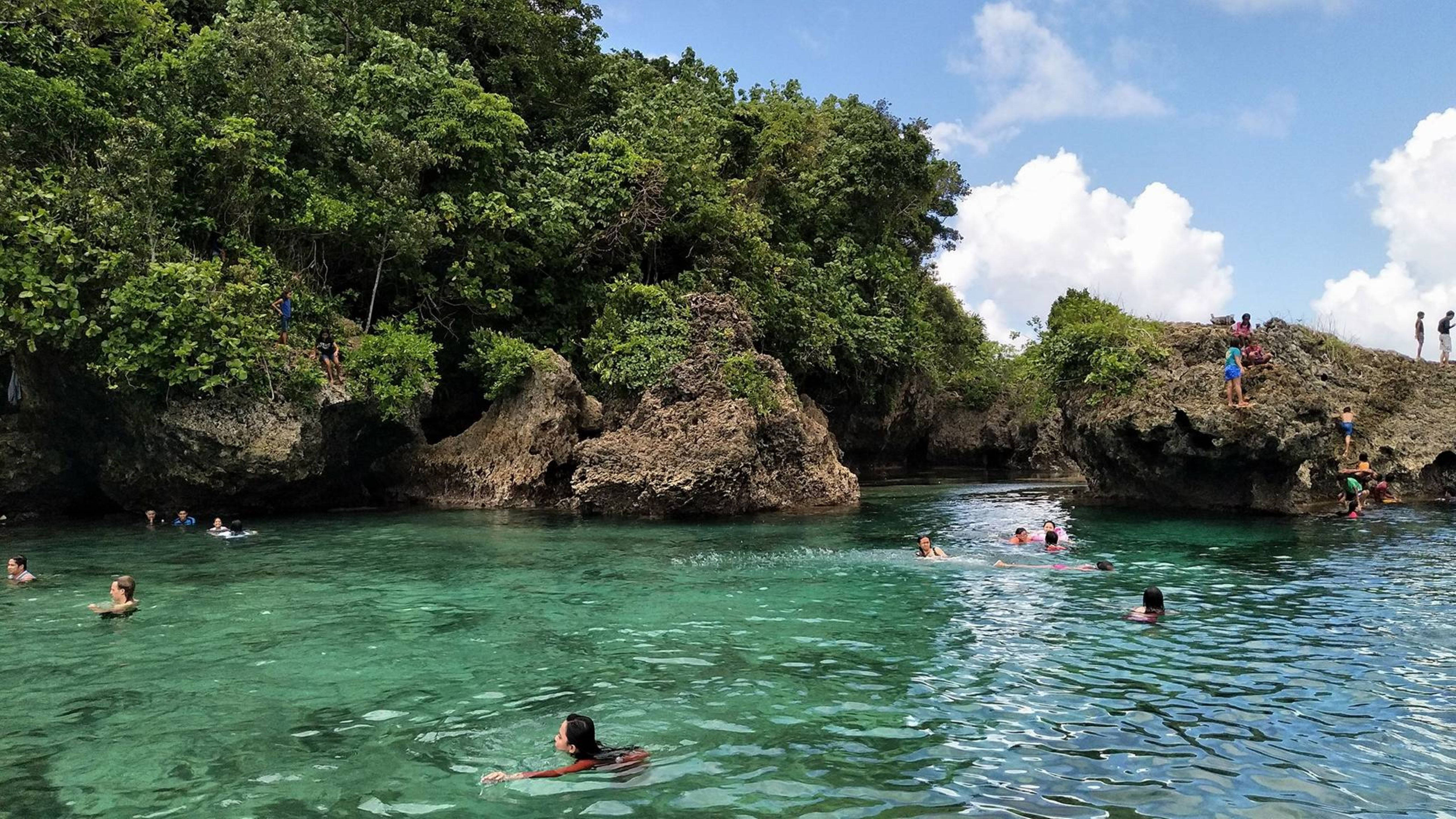 Siargao 桌布 壁紙 — 人們在清澈的碧綠海水中游泳和玩耍，周圍是茂密的綠色懸崖，頭頂是明亮的藍天。