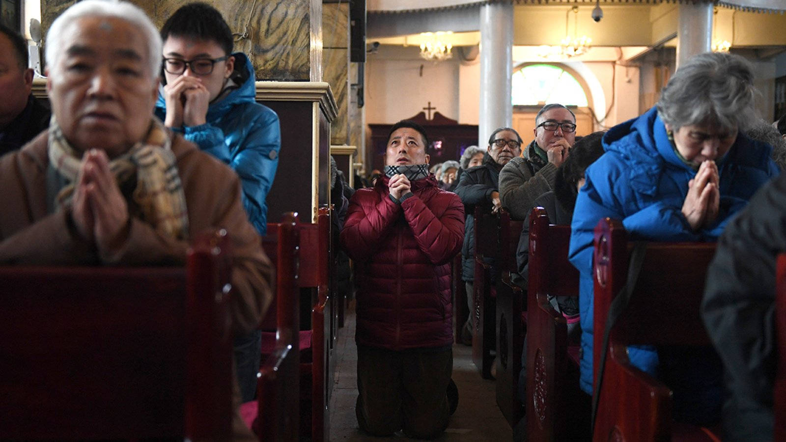 教堂 桌布 壁紙 — 人們在溫暖照明的教堂內跪著祈禱，教堂有木製長椅和拱形窗戶。