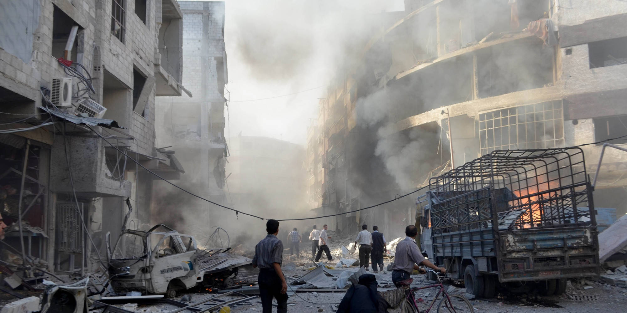 Syria 桌布 壁紙 — 人們在煙霧彌漫的戰區中穿行，周圍是毀壞的建築和燃燒的殘骸。
