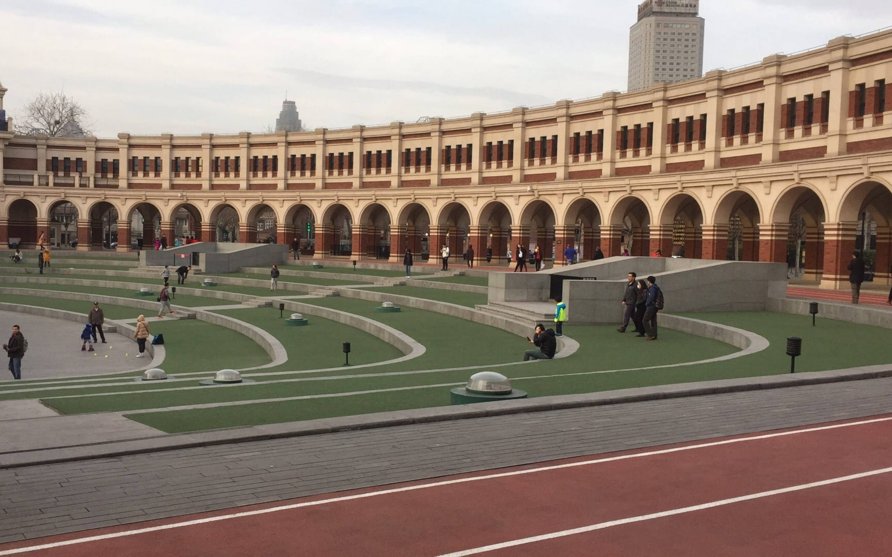 Tianjin 桌布 壁紙 — 人們在現代化的綠色梯田廣場上漫步和玩耍，廣場上方是宏偉的拱形柱廊，前景是紅色跑道。