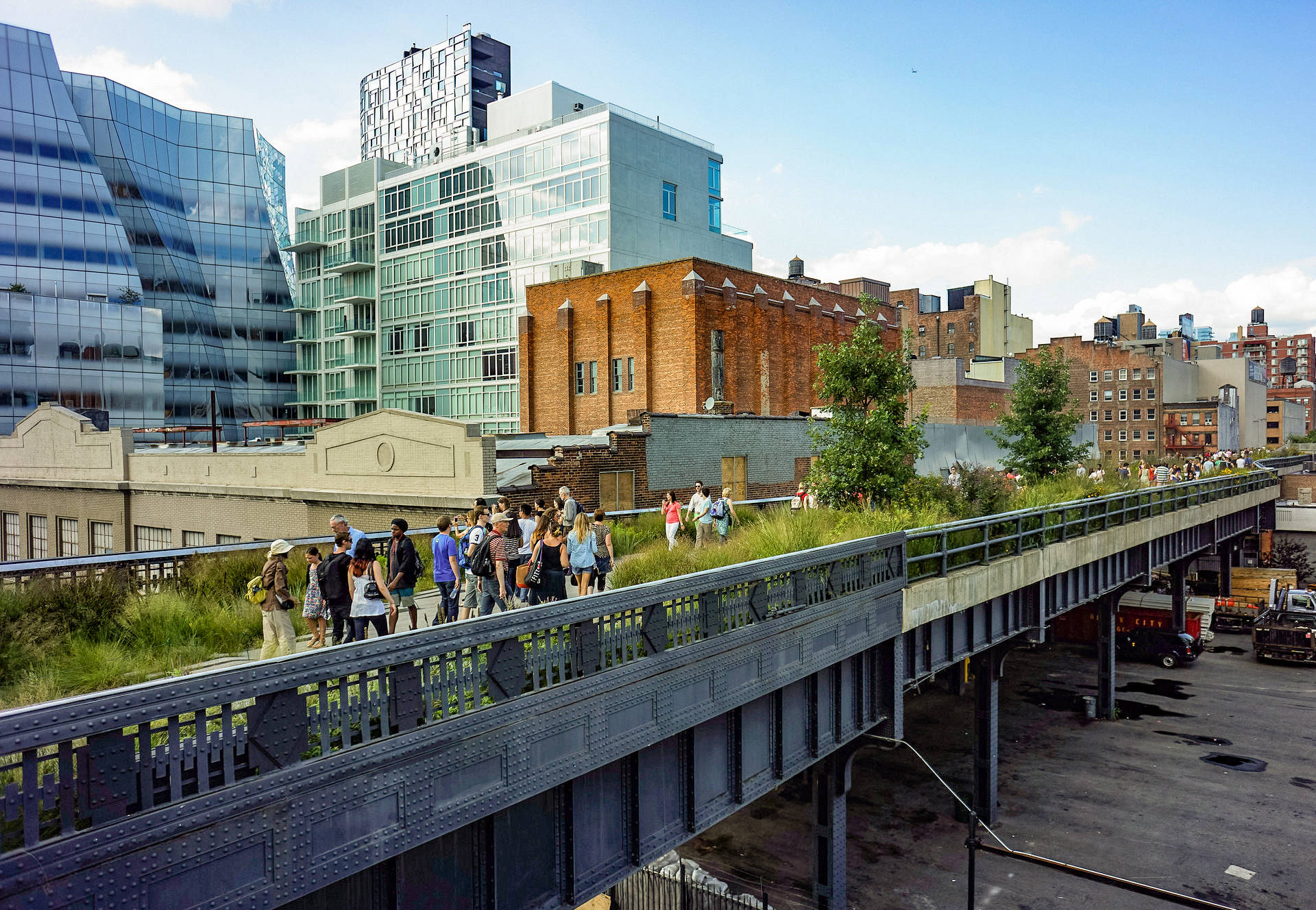 New York 桌布 壁紙 — 人們在現代建築和磚砌建築的城市街道上方，沿著一條長滿草的高架步道漫步，天空湛藍清澈。
