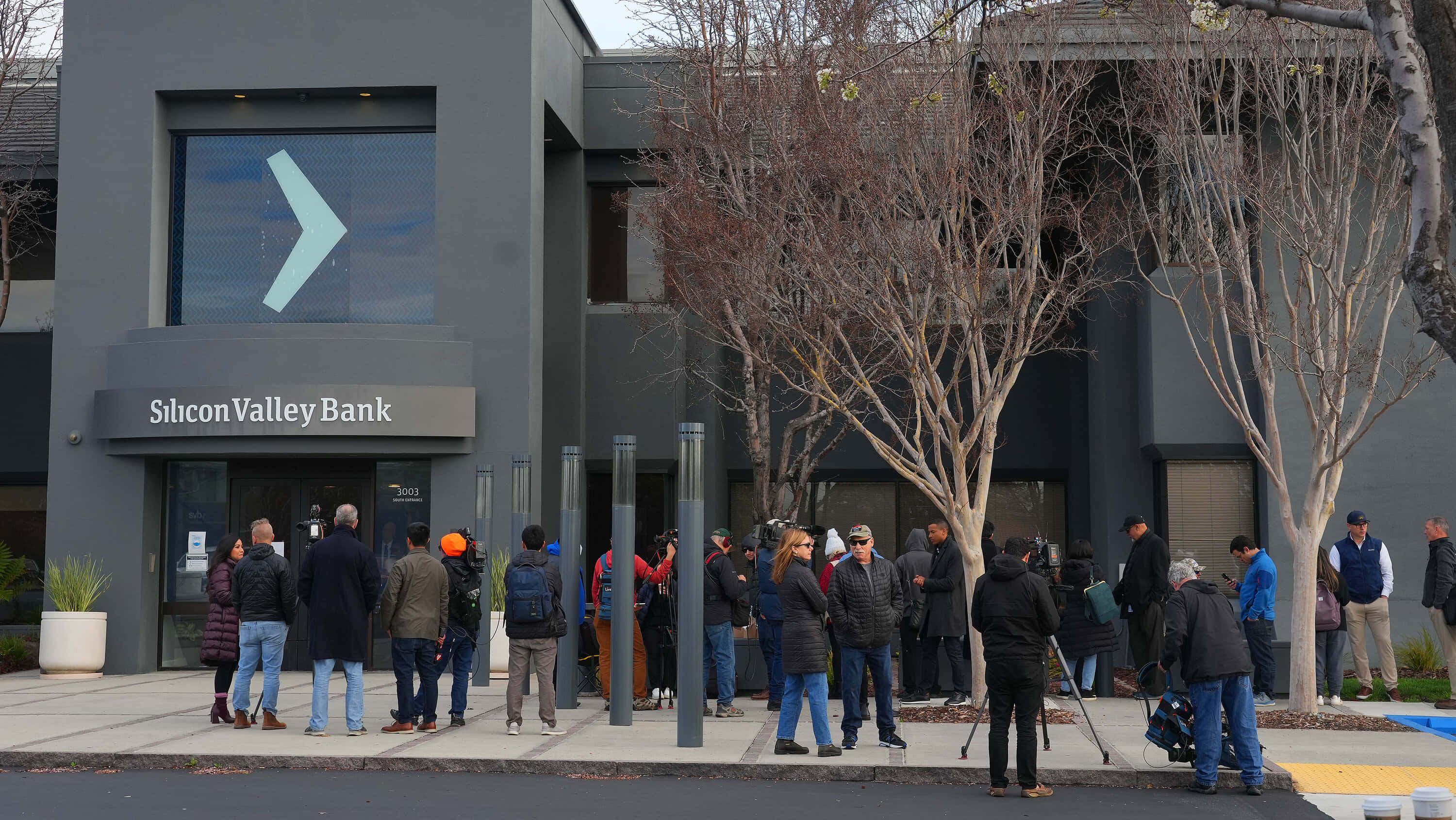 Bank 桌布 壁紙 — 人們在現代灰色矽谷銀行大樓外排隊，附近有光禿禿的樹木。