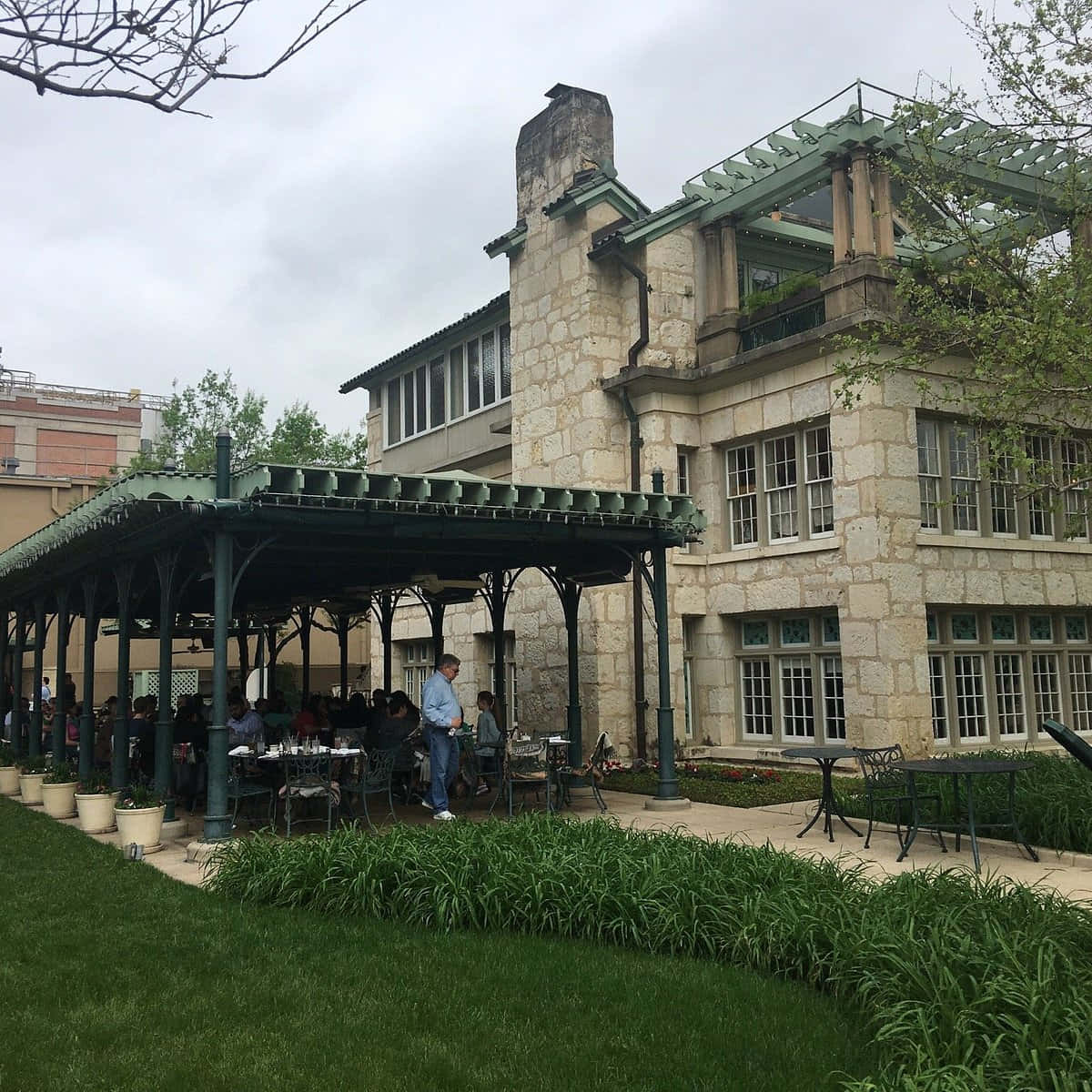 San Antonio 桌布 壁紙 — 人們在石建築旁邊的綠色藤架下用餐，周圍有茂密的綠植和陰天。