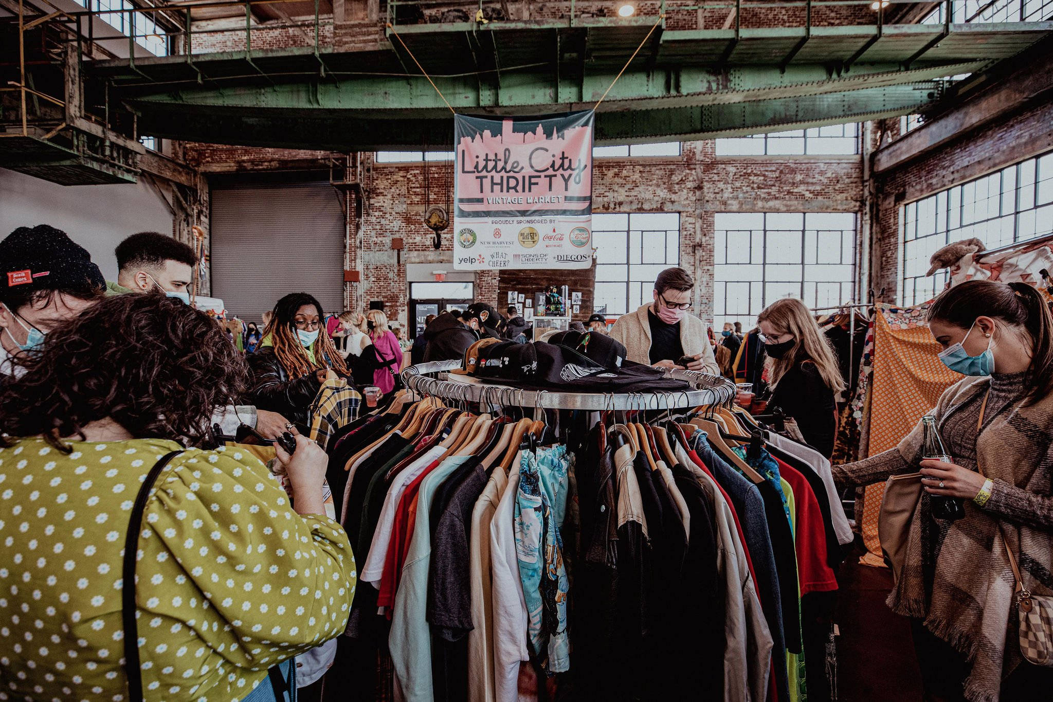 Clothes 桌布 壁紙 — 人們在繁忙的室內二手市場中，在裸露的磚牆和鋼樑下瀏覽色彩繽紛的復古服裝。
