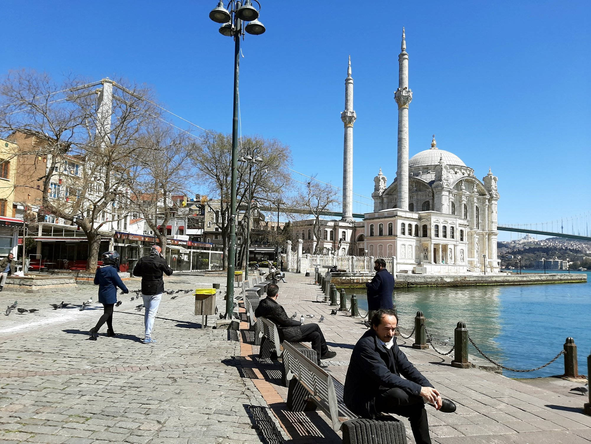 Istanbul 桌布 壁紙 — 人們在蔚藍天空下，於海濱長椅放鬆，旁邊有宏偉的白色清真寺和橋樑。