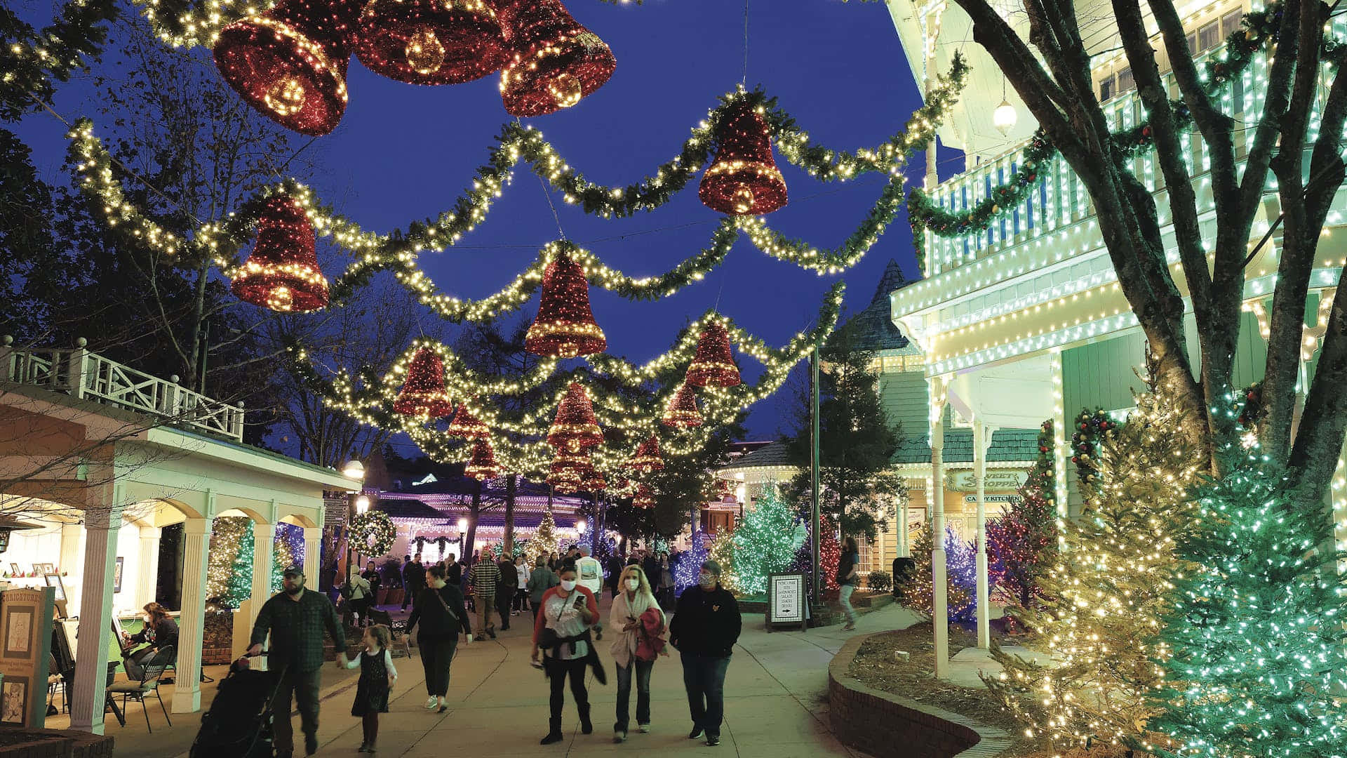 Christmas Village 桌布 壁紙 — 人們在裝飾過的城鎮廣場上，在閃爍的紅色鈴鐺和串燈下漫步，度過一個節日假期夜晚。