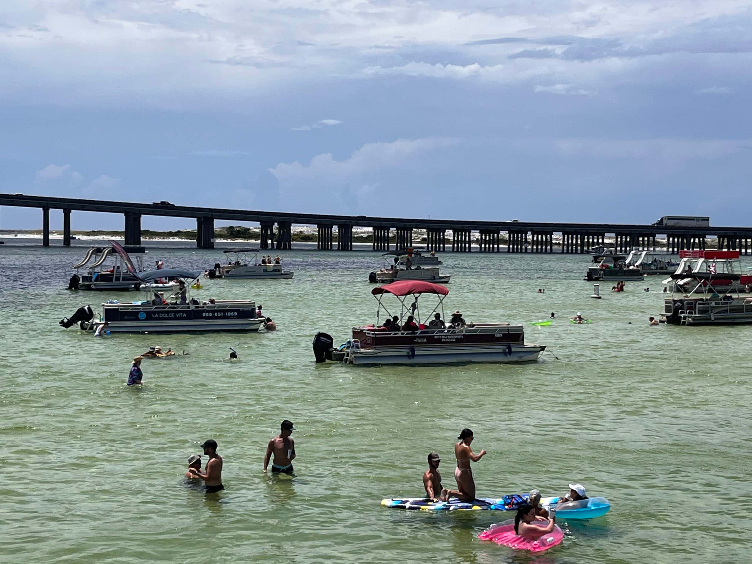 Crab Island 桌布 壁紙 — 人們在陰天之下，靠近一條長橋，在淺綠色水中游泳和漂浮在木筏上，附近有船隻。