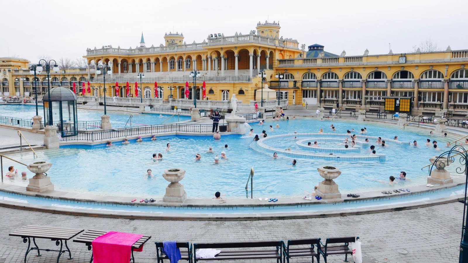 Vacation 桌布 壁紙 — 人們在陰天時在一個被黃色新古典主義建築包圍的大型戶外溫泉池中放鬆。