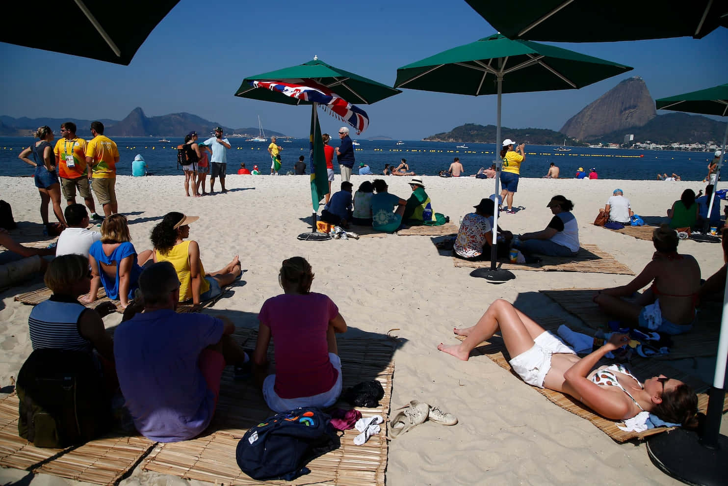 Brazilian Beach 桌布 壁紙 — 人們在陽光明媚的海灘上，在綠色遮陽傘下放鬆，背景是糖麵包山和海洋。