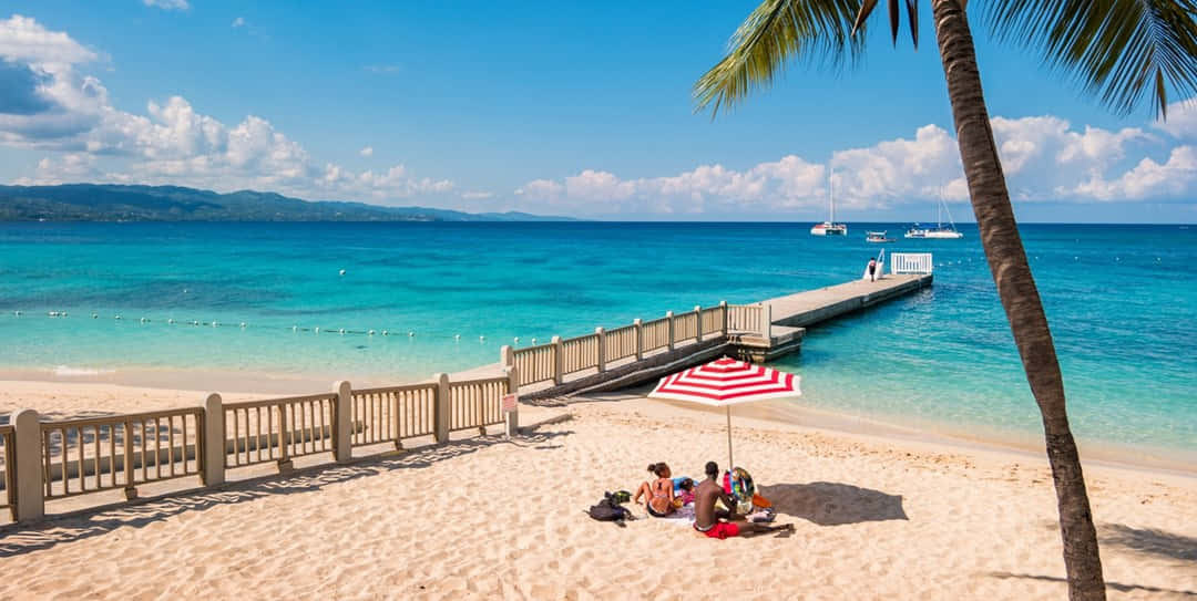 Jamaican Beach 桌布 壁紙 — 人們在陽光明媚的熱帶海灘上，在紅白條紋的遮陽傘下放鬆，海水呈碧綠色，木製碼頭延伸入海。
