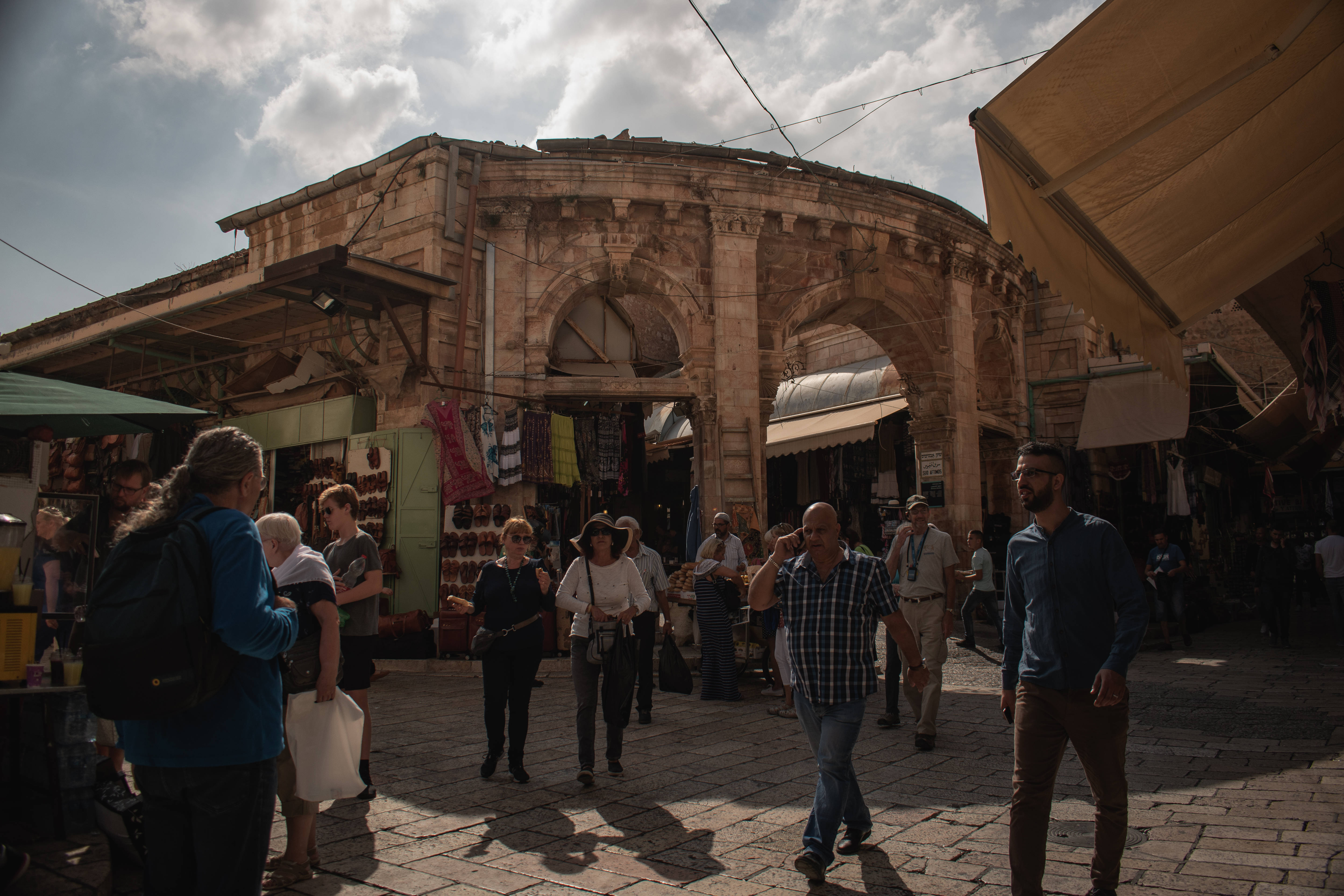 Israel 桌布 壁紙 — 人們在陽光普照的歷史市場中漫步，市場有石拱門和色彩繽紛的攤位，天空局部多雲。