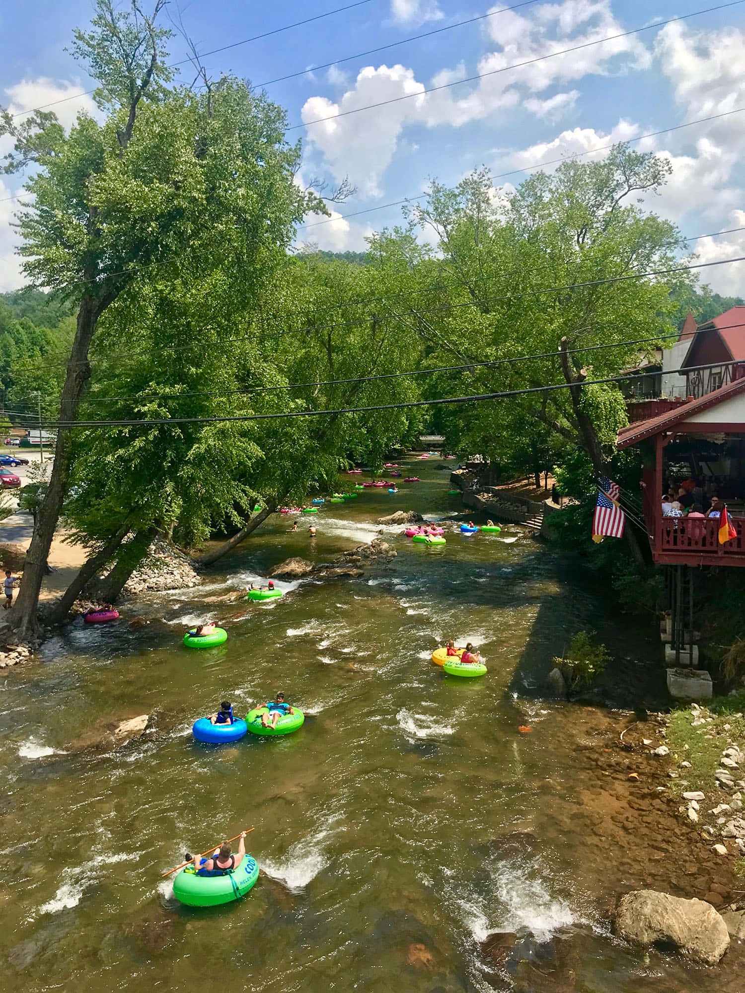 夏天 桌布 壁紙 — 人們在陽光照耀的河流上乘坐色彩繽紛的浮圈漂流，周圍是茂密的綠樹和一座木製河邊建築。