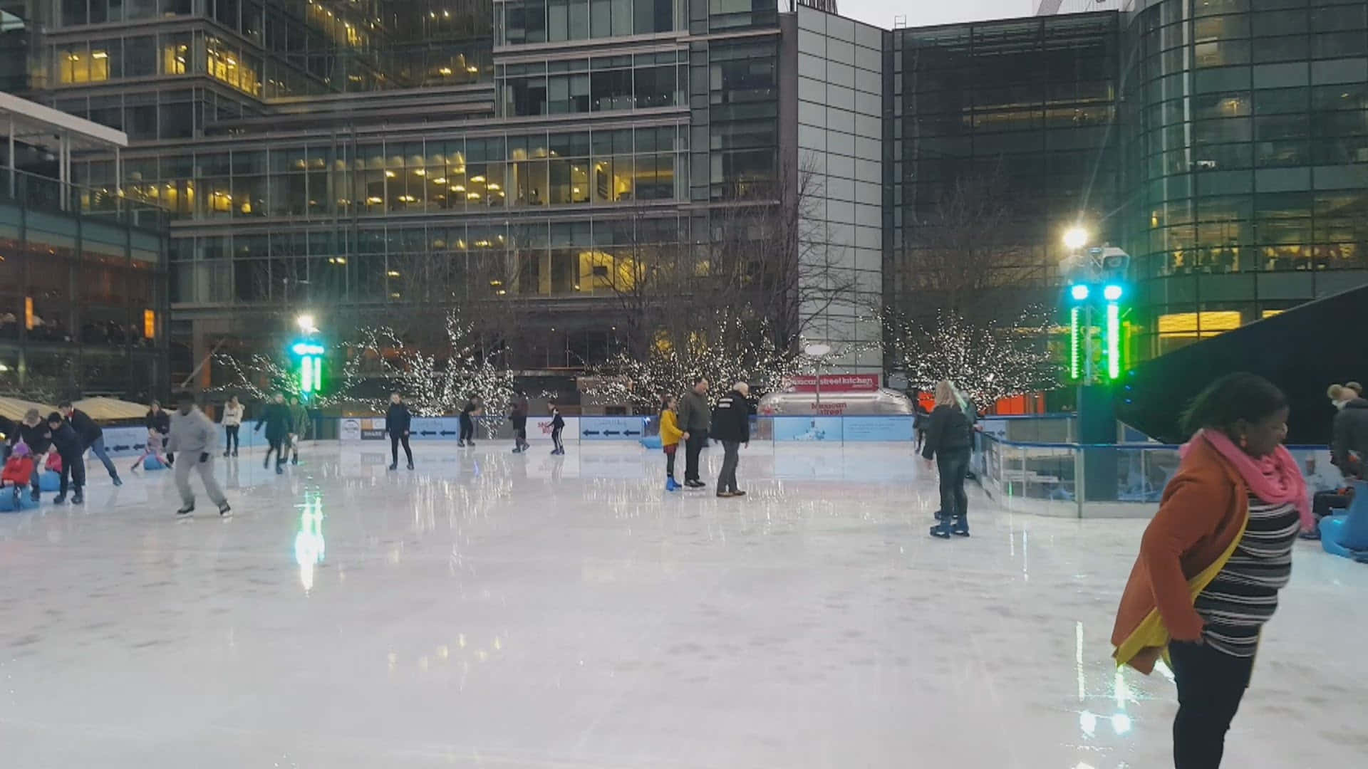 Ice Rink 桌布 壁紙 — 人們在黃昏時分，在現代玻璃建築環繞的閃爍戶外溜冰場上滑行。