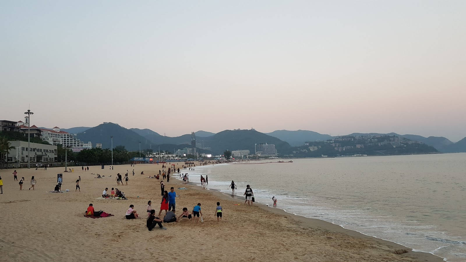 Shenzhen 桌布 壁紙 — 人們在黃昏時分沿著沙灘放鬆和玩耍，溫柔的海浪和遠處的山脈在柔和的粉紅色和藍色天空下。